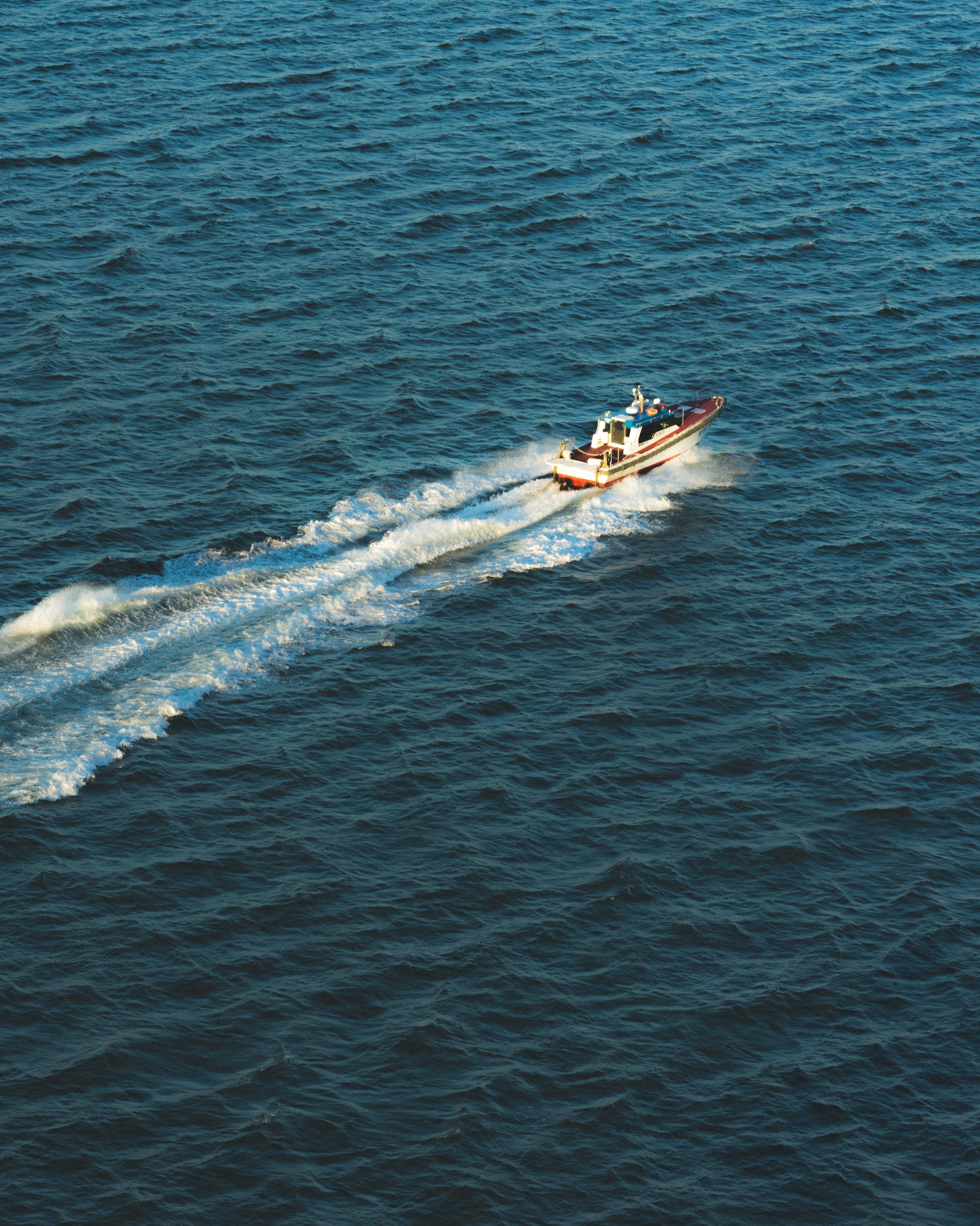 A small speedboat moves swiftly across deep blue ocean waters, leaving a trail of white wake behind it. The boat, positioned slightly off-center to the right, is angled diagonally toward the top of the image. The water around the boat is textured with small waves and ripples.