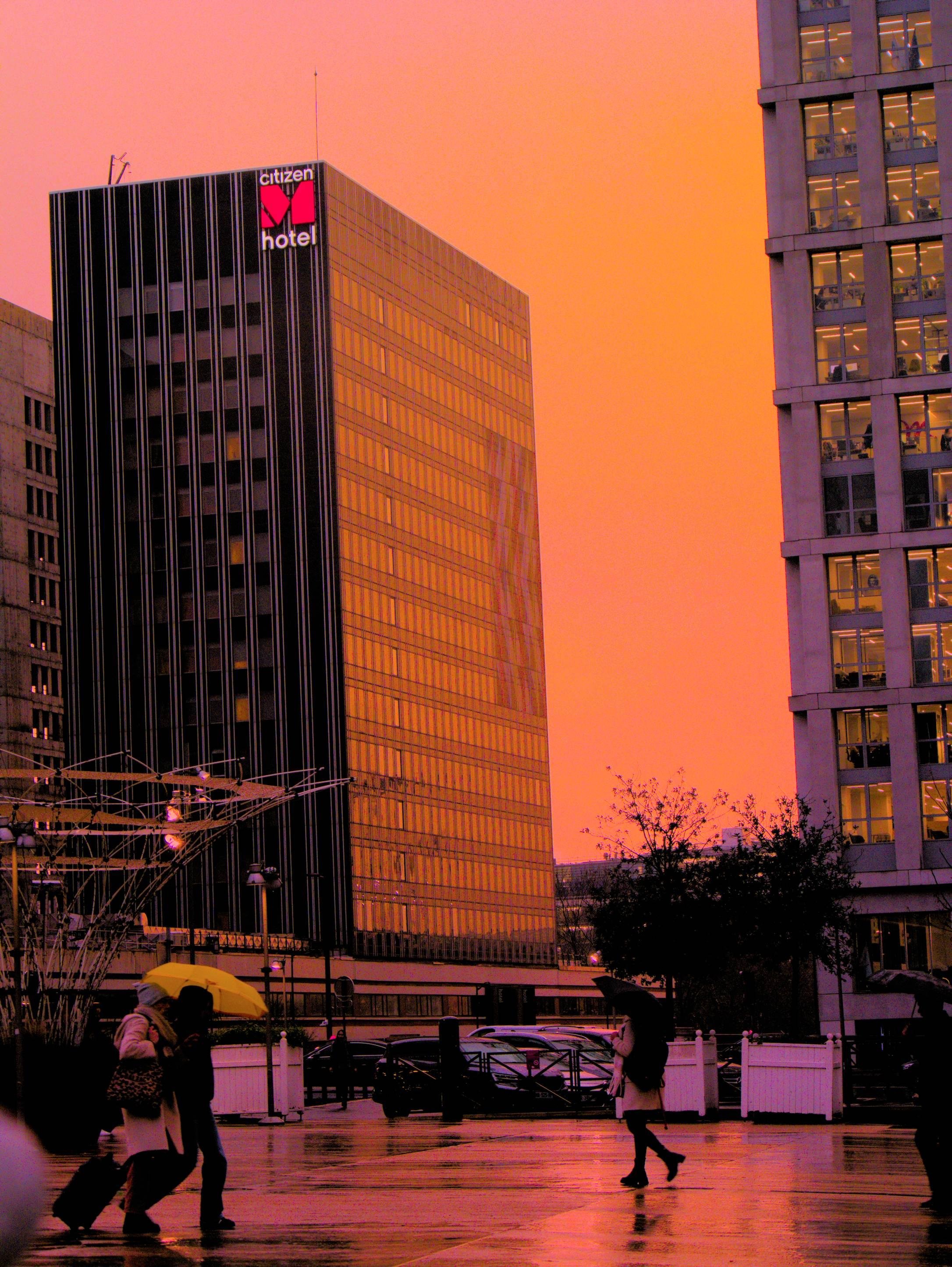 La photographie représente une scène urbaine au coucher du soleil, avec un bâtiment portant le logo « citizenM hotel ». Le ciel a une teinte orange chaude et des personnes marchent au premier plan, certaines portant des parapluies. Les bâtiments reflètent la couleur du coucher de soleil, créant une atmosphère vibrante.