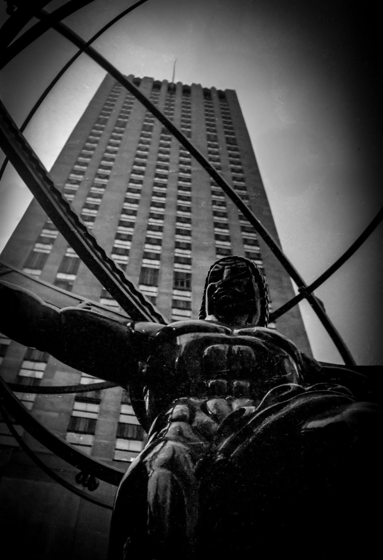 Large bronze sculpture of a muscular figure holding a spherical armillary, viewed from below. Behind the sculpture, a tall skyscraper with numerous windows stretches into the sky. The image is in black and white, enhancing the contrast between the dark statue and the lighter building.