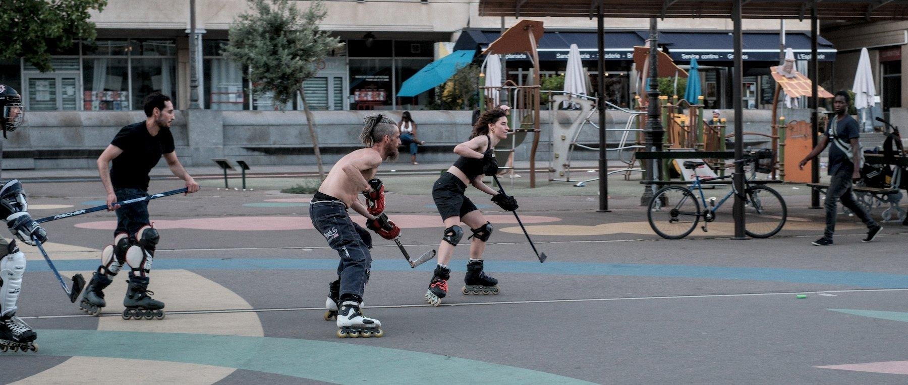 Photo horizontale. Sur une place piétonne en bitume, 3 hommes et une femme sur roller et des protections tantôt au genoux, tantôt au tibia, tantôt aux mains, attendent de pieds ferme le palet vert