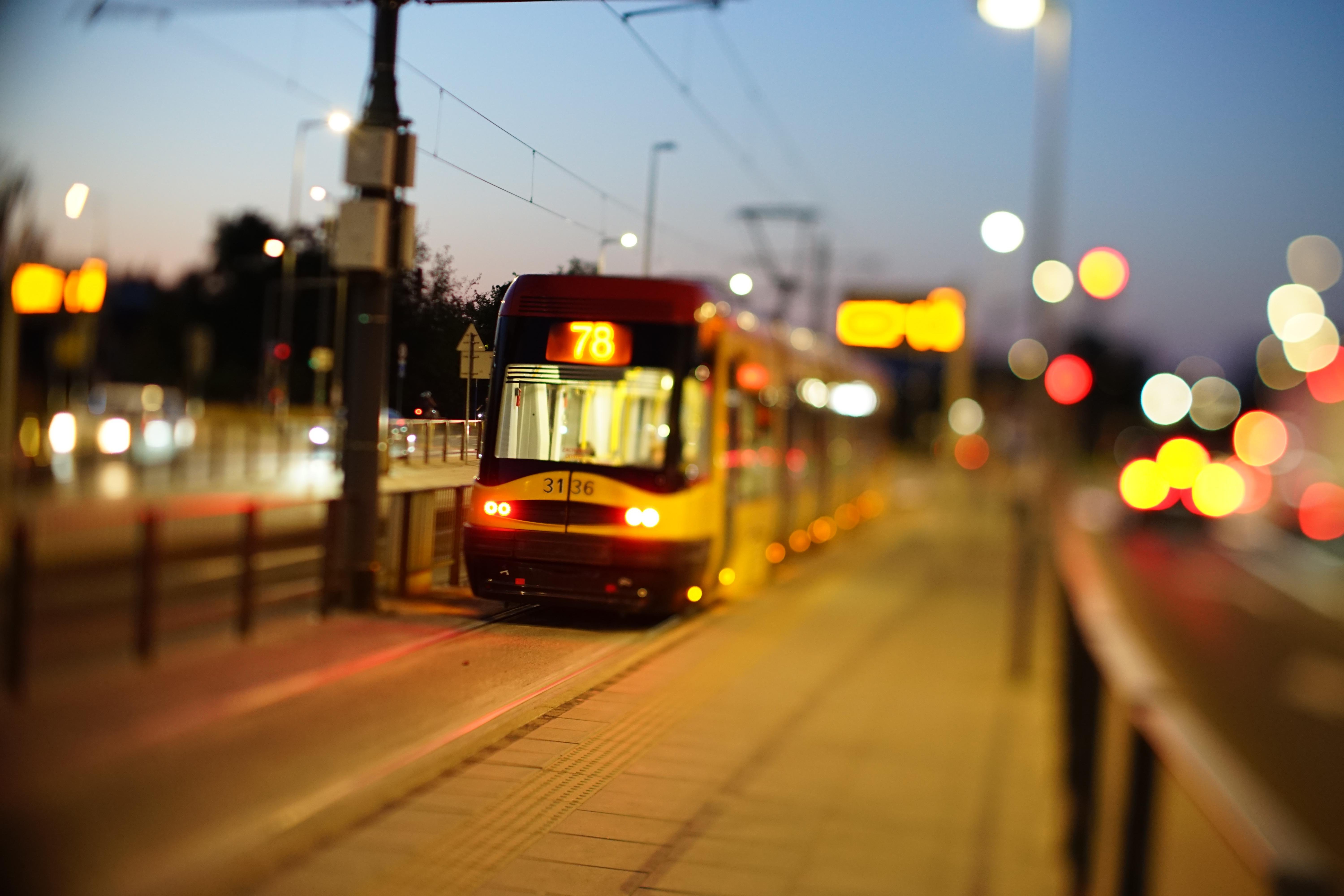 A yellow and red tram with the route number 78 illuminated on its front travels along a city street during dusk. The background and foreground feature blurred streetlights and buildings, creating a bokeh effect. Overhead wires and a platform are visible, with the sky transitioning from blue to darker shades as evening approaches.