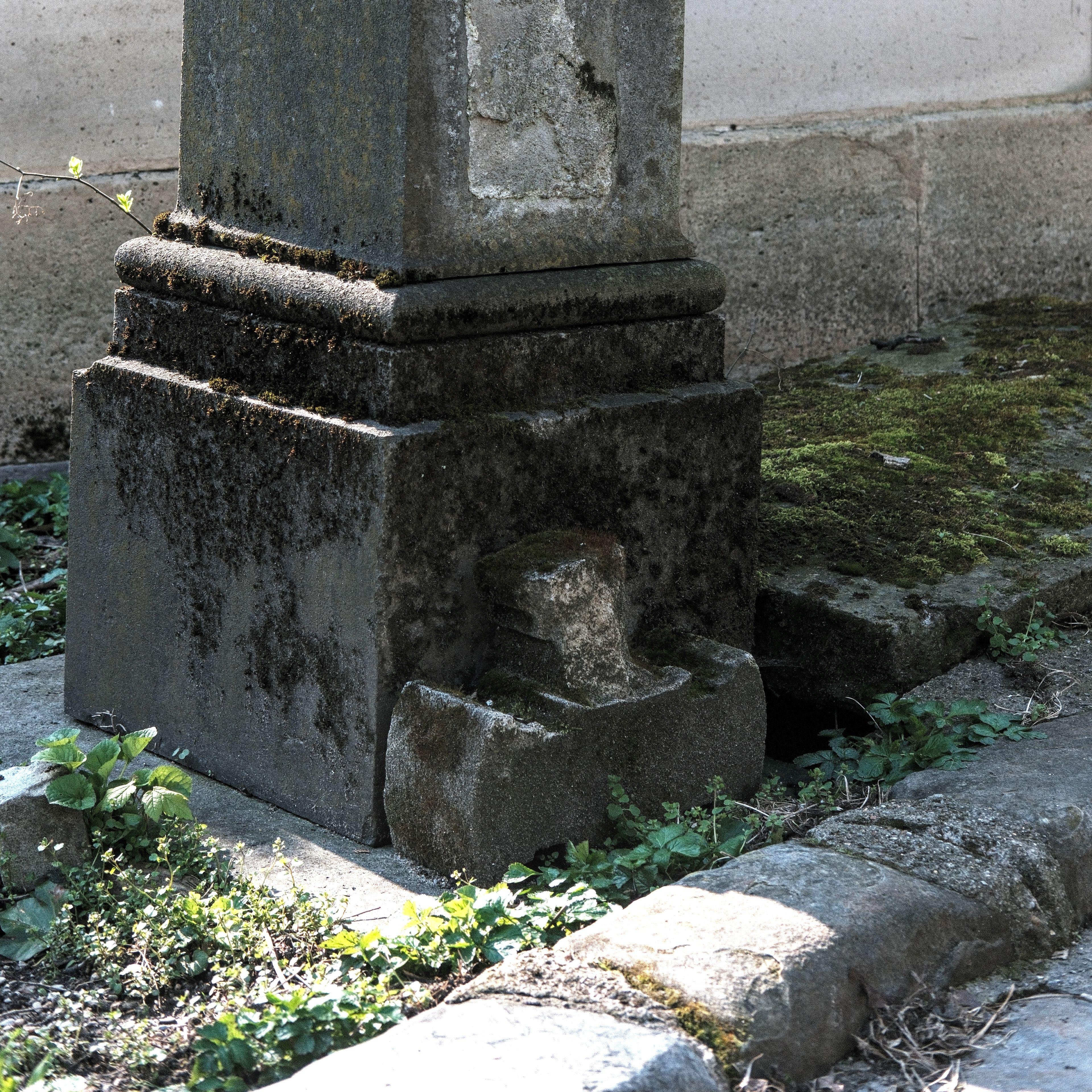 Photo carrée. Au pieds d’une grande stèle, un morceau de croix brisé repose contre le socle. Le tout est couvert de mousse sombre.
