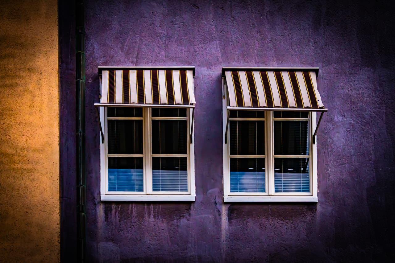 Two adjacent windows with striped awnings on a purple wall. Each window has white frames and closed blinds. An orange wall is partially visible on the left side of the image.