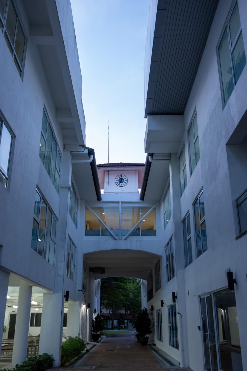 Narrow alleyway bordered by tall, white buildings with numerous windows on each side. Overhead, a bridge connects the buildings, featuring a prominent circular clock. The pathway below is paved with bricks, leading to an area with trees in the background. The sky is a soft blue, suggesting early evening or morning light.