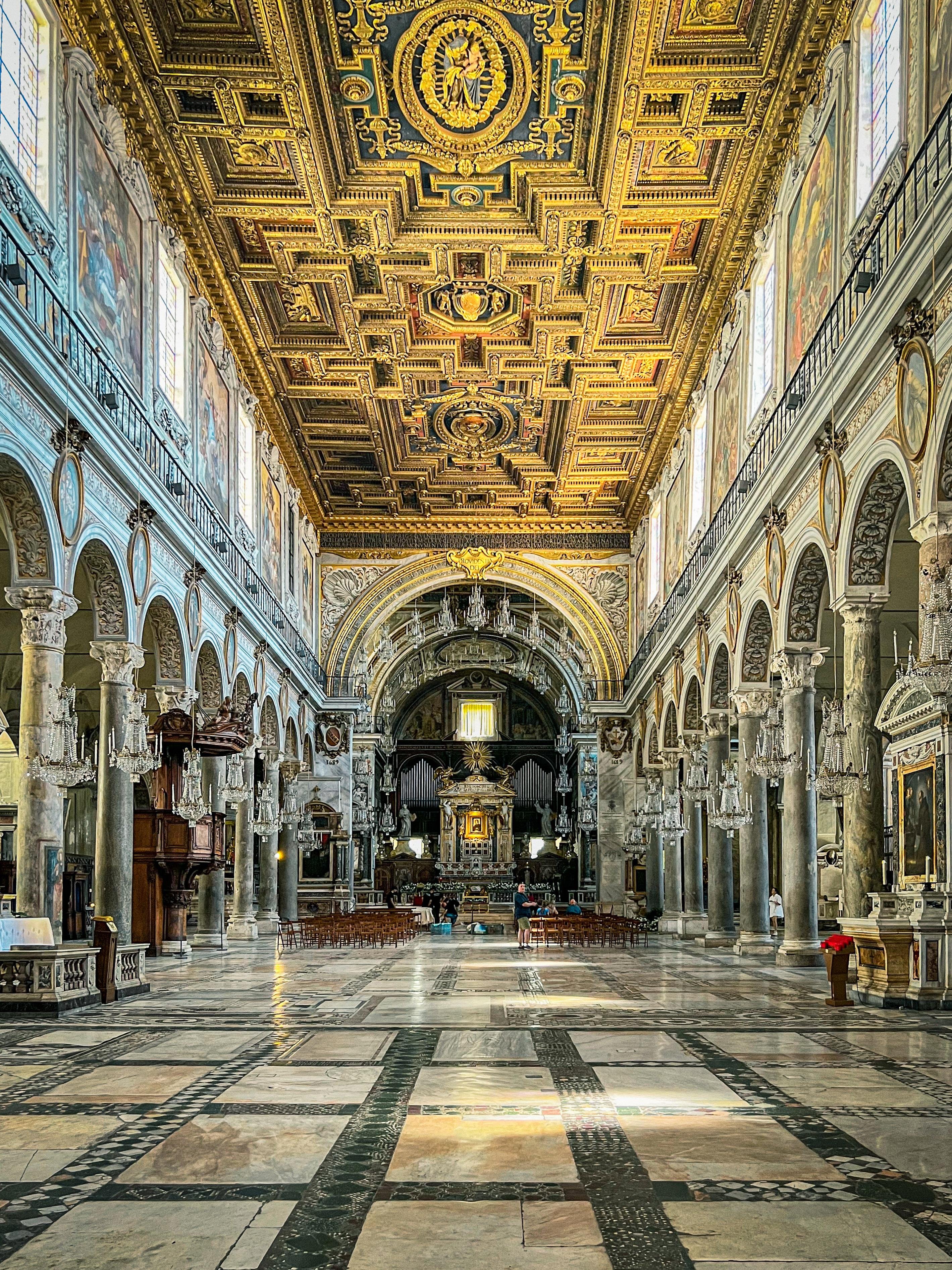 Interno di una grande chiesa. Il soffitto è ornato con intricati disegni dorati e opere d'arte elaborate. L'interno presenta grandi colonne e archi su entrambi i lati, con lampadari appesi al soffitto. Il pavimento è in pietra lucida e lo stile architettonico complessivo è riccamente dettagliato, tipico dei design classici o rinascimentali. All'estremità opposta, sembra esserci un altare o un punto focale centrale, e lo spazio è riempito di luce naturale che entra attraverso grandi finestre.
