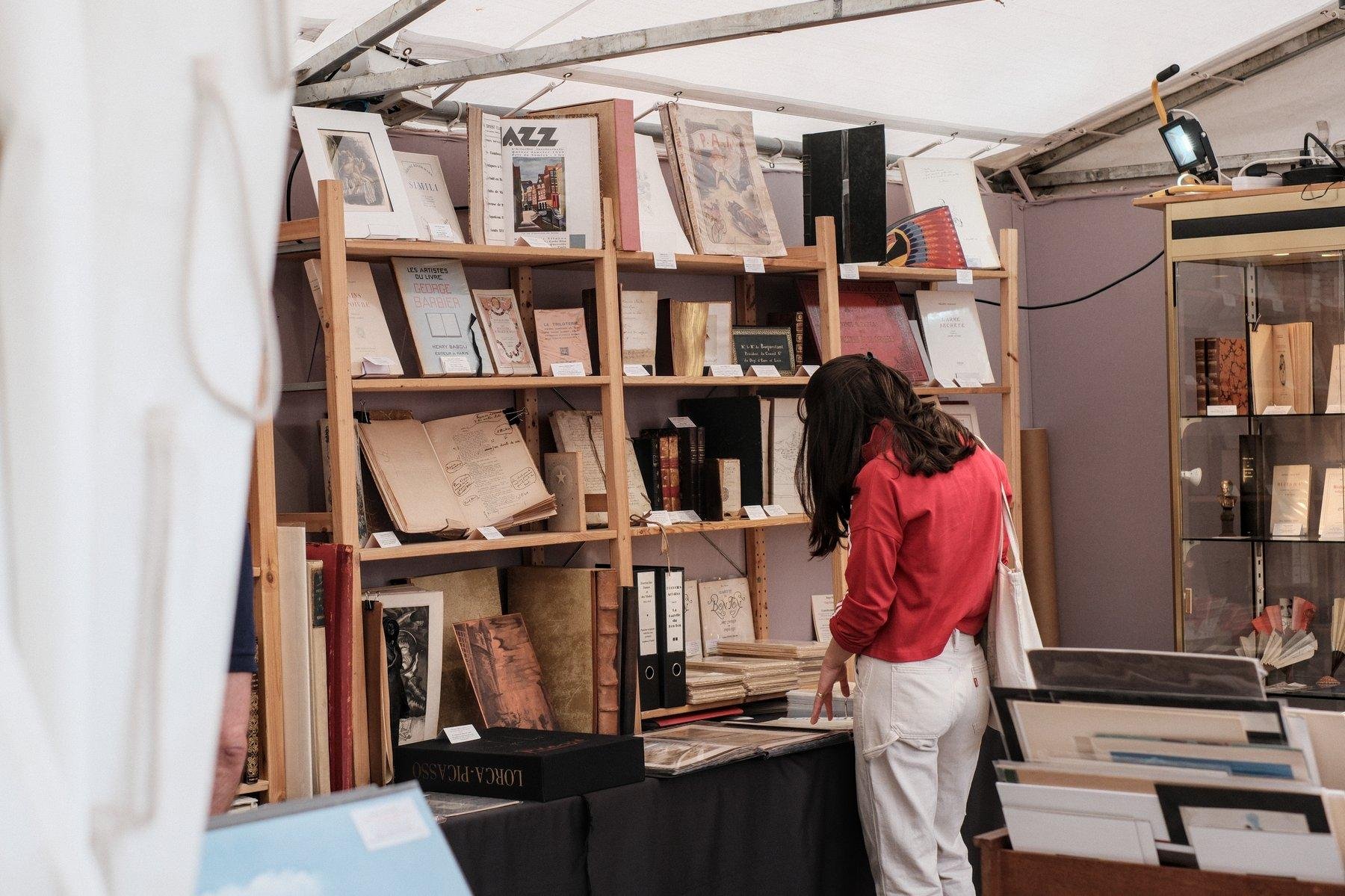 Photo horizontale. Sous une tente de toile blanc, une librairie temporaire est installée. Une jeune femme brune au cheveux long, avec un chemisier rouge et un pantalon blanc, de dos, est en train de feuilleter un gros classeur aux feuillets plastiques