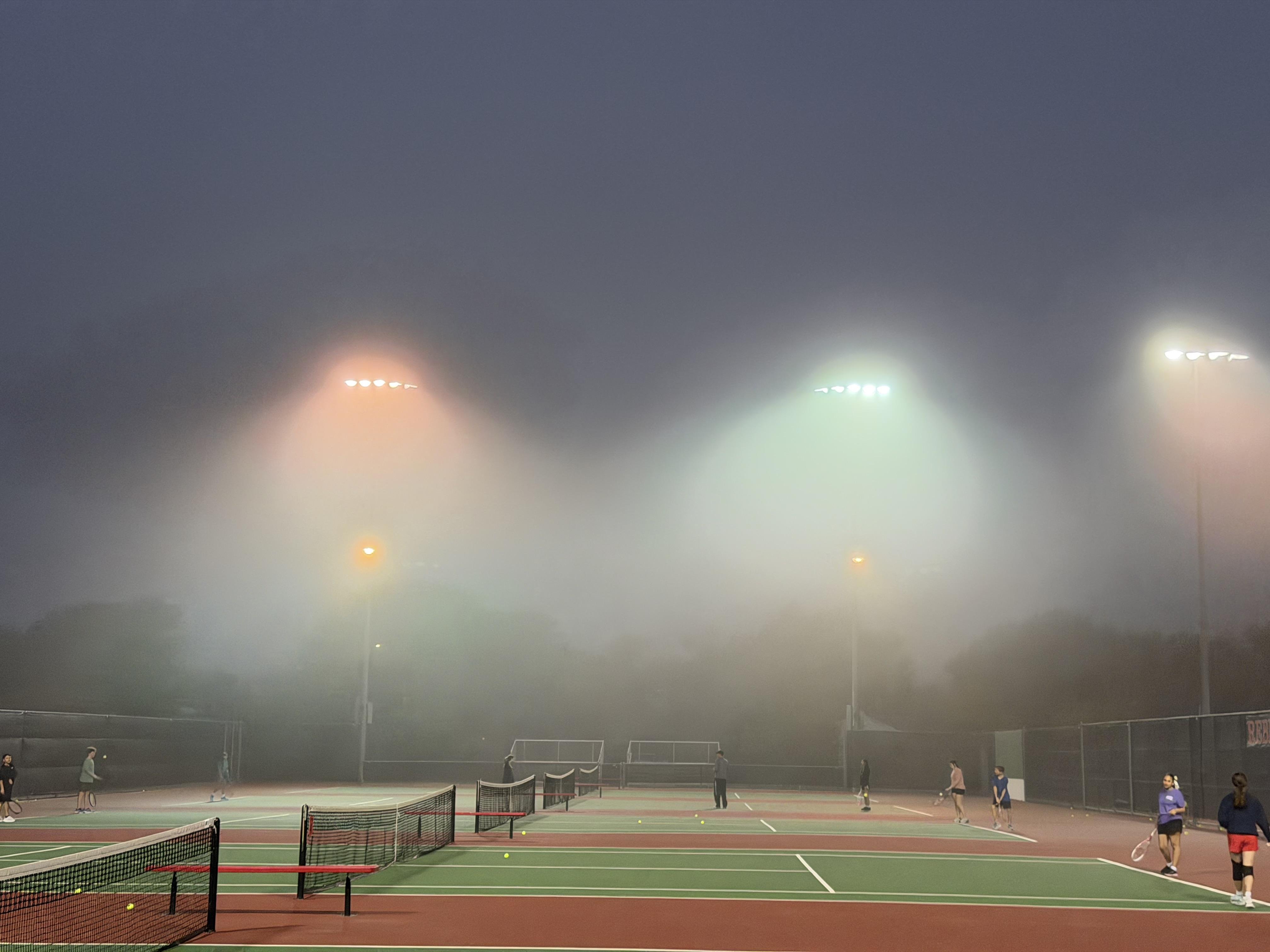 Foggy early morning scene on an outdoor tennis court with multiple players. Bright floodlights illuminate the area, casting a soft glow through the mist. Several tennis nets are visible, and players are scattered across the courts, some holding rackets. Trees and fencing are faintly seen in the background, partially obscured by fog.