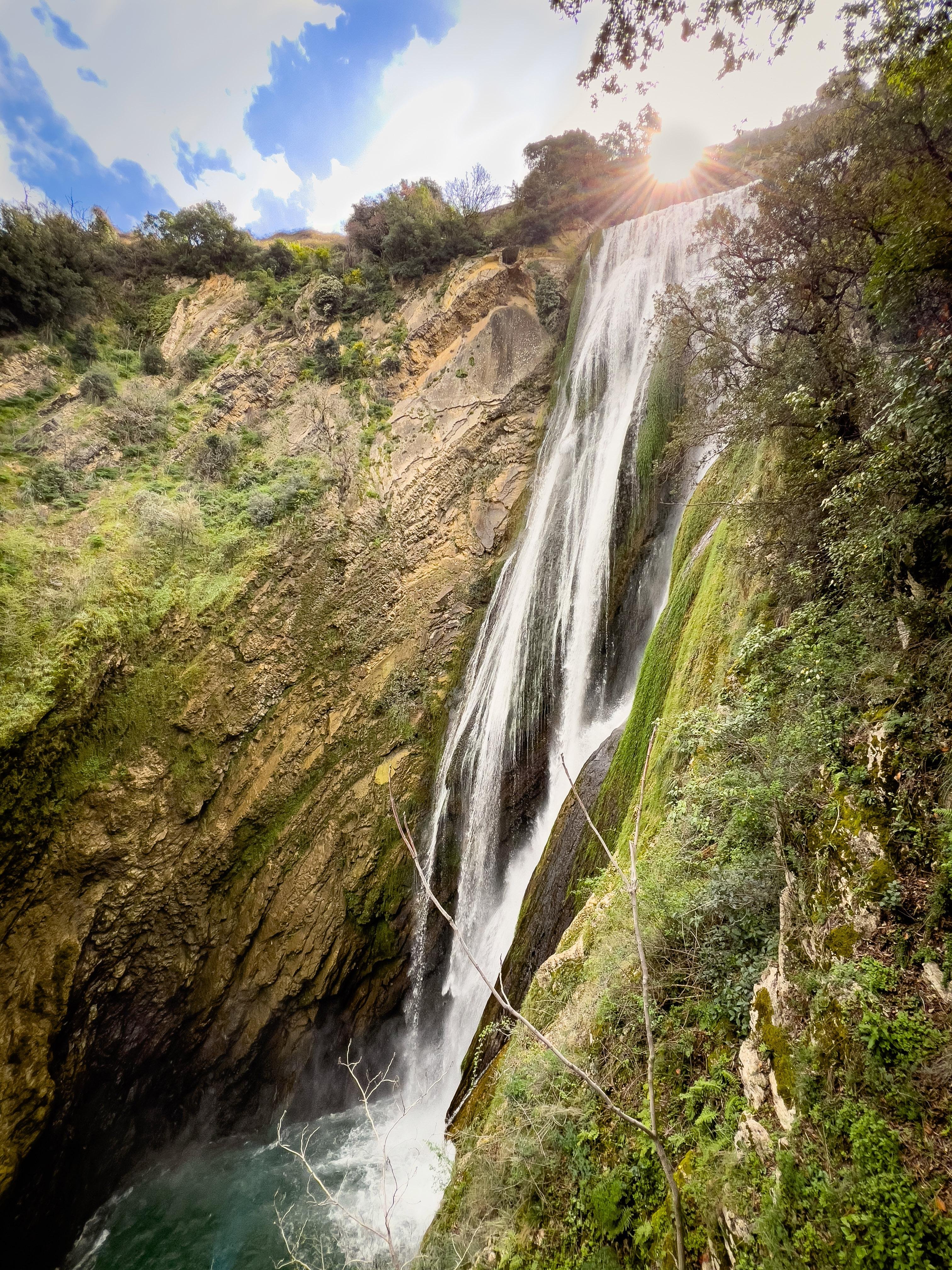Una cascata che scende lungo una scogliera rocciosa in una piscina sottostante. L'area circostante è rigogliosa di vegetazione, e il sole è parzialmente visibile in alto, irradiando una calda luce sulla scena.
