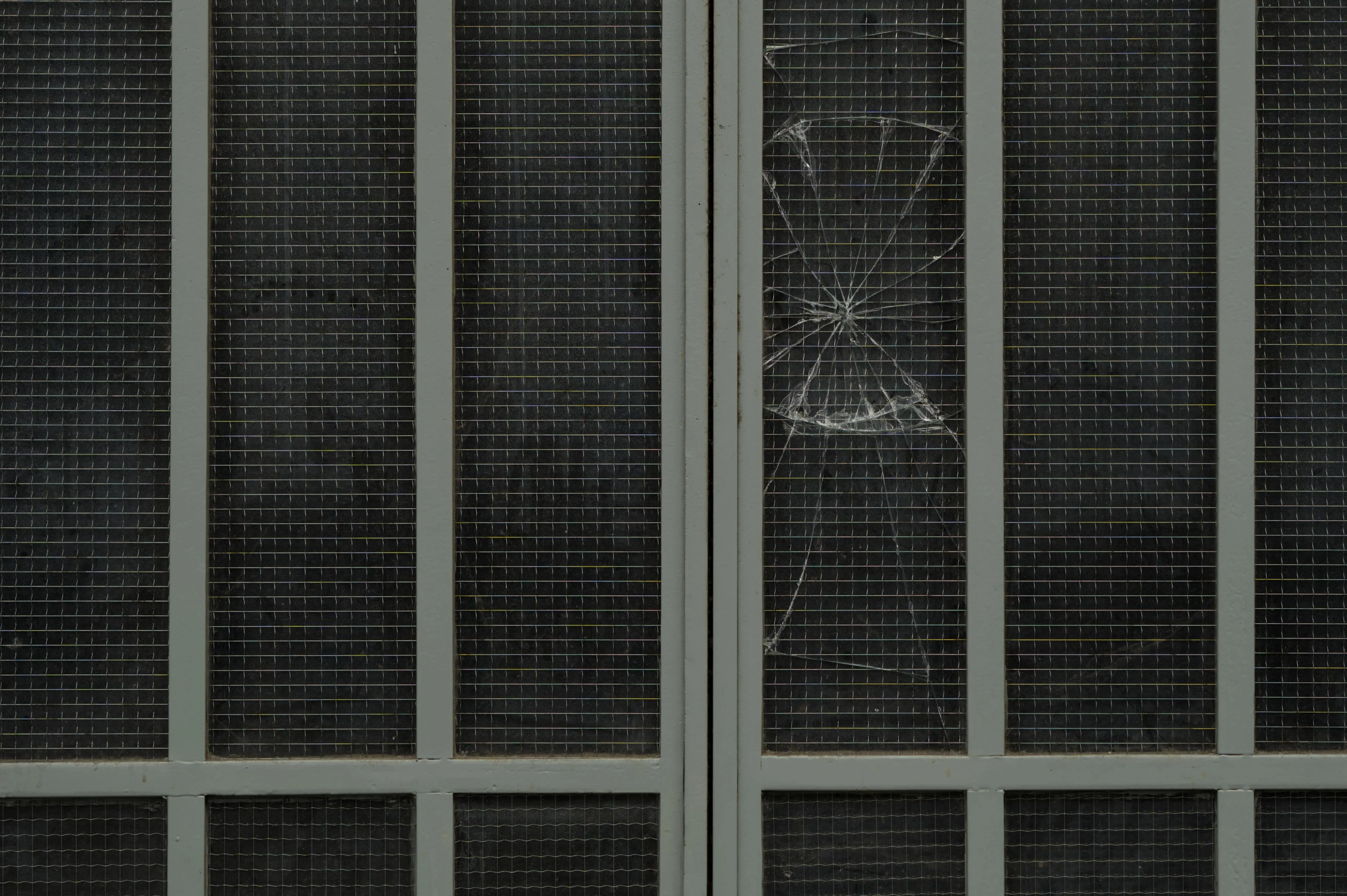 Close-up photograph of a grate window backed by a dark, opaque glass surface surrounded with metallic, light green window bars. One of the window panes has its glass broken, forming cracks centered on a single point. The mesh over the glass of window panes reflects light with rainbow colors. The overall photograph has a minimalist style with repeated elements of window panes and the grate mesh.