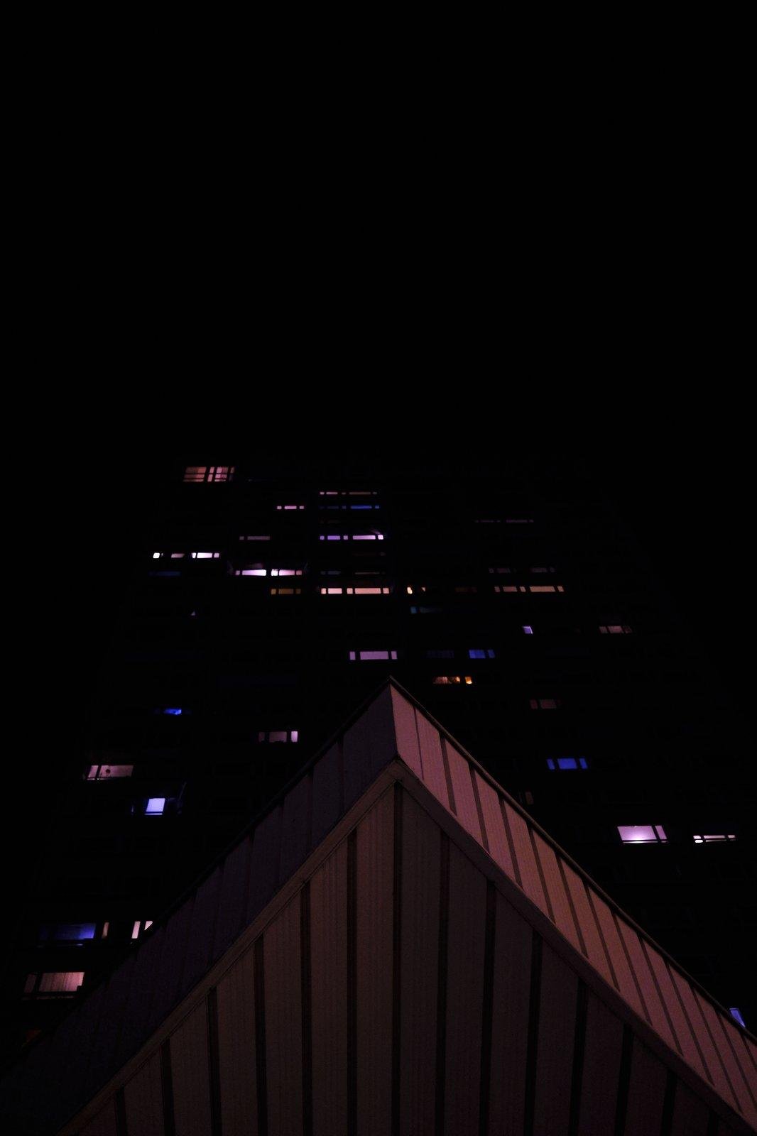 Photo format portrait, de nuit, en contre-plongée, avec sur le tiers du bas, un triangle d’un bout de toit, et derrière, quelques lumières émanant de fenêtres d’un grand immeuble invisible dans la nuit