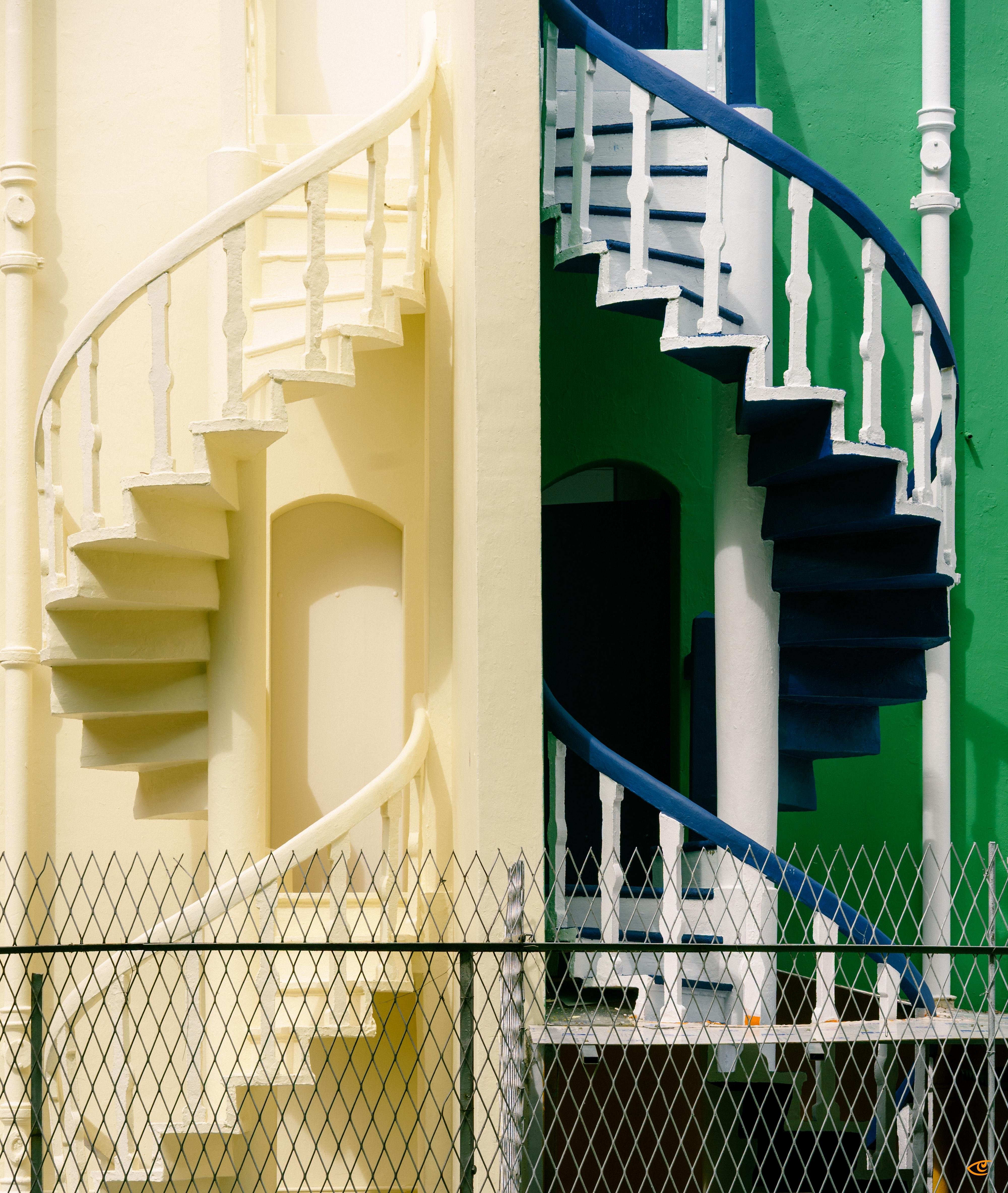 Two adjacent spiral staircases separated by a vertical wall: left staircase painted cream with white railing, right staircase painted dark blue with white railing against a green wall, with a chain-link fence along the bottom foreground.