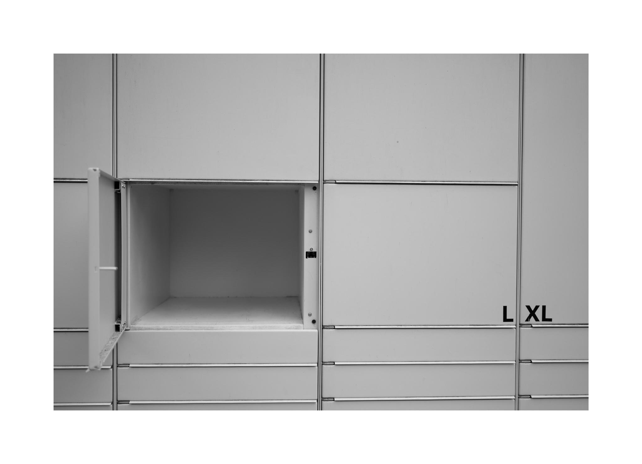 hwarz-Weiß Nahaufnahme einer Packstation mit geöffnetem Fach. Nur die Umrandung der Fächer sind sichtbar. Im rechten Bildrand sind die Beschriftungen L und XL zu lesen.