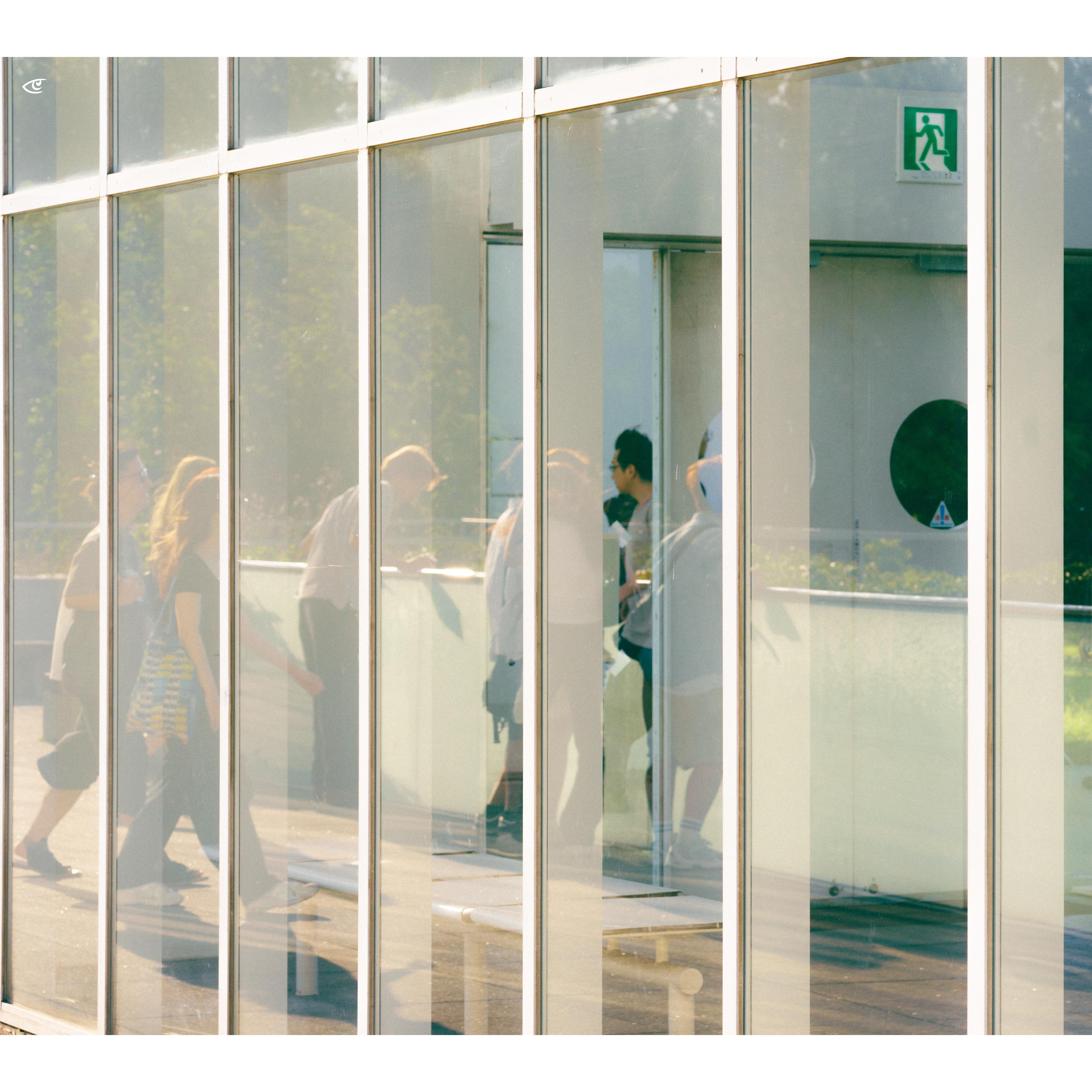 People walking and standing inside a glass-walled corridor, seen through tall vertical window panes with reflections; green emergency exit sign near the top right above a door.