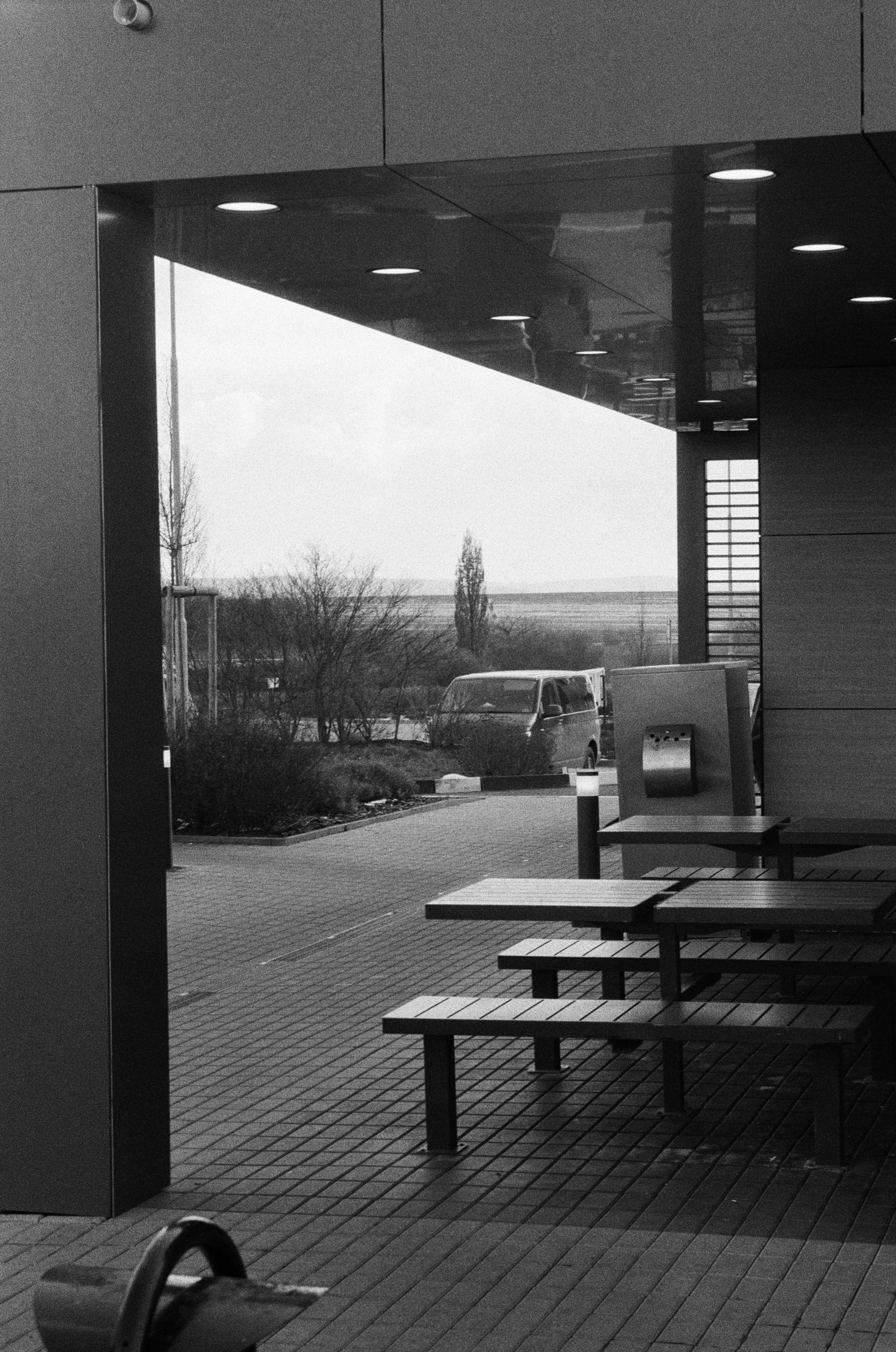 The black and white image shows an outdoor seating area with benches and tables. There is a modern structure with a roof over it, and some recessed ceiling lights are visible. In the background, there are trees and a parked vehicle. The setting appears to be an outdoor public or commercial space, possibly adjacent to a building.