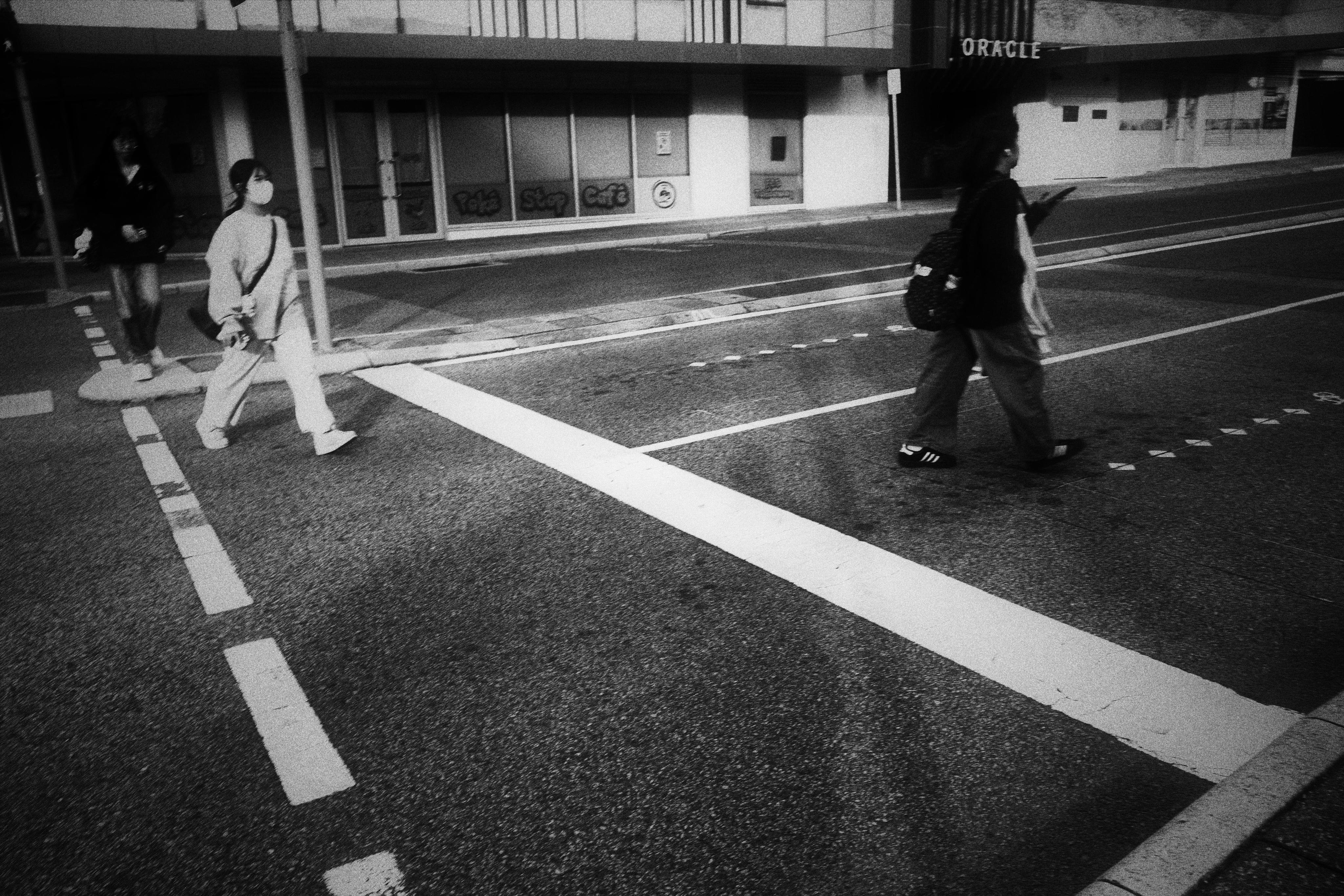 Black-and-white street scene with three pedestrians crossing a marked crosswalk; one person walks across the right side of the road while two step off the curb on the left, with storefront windows and a sidewalk in the background.