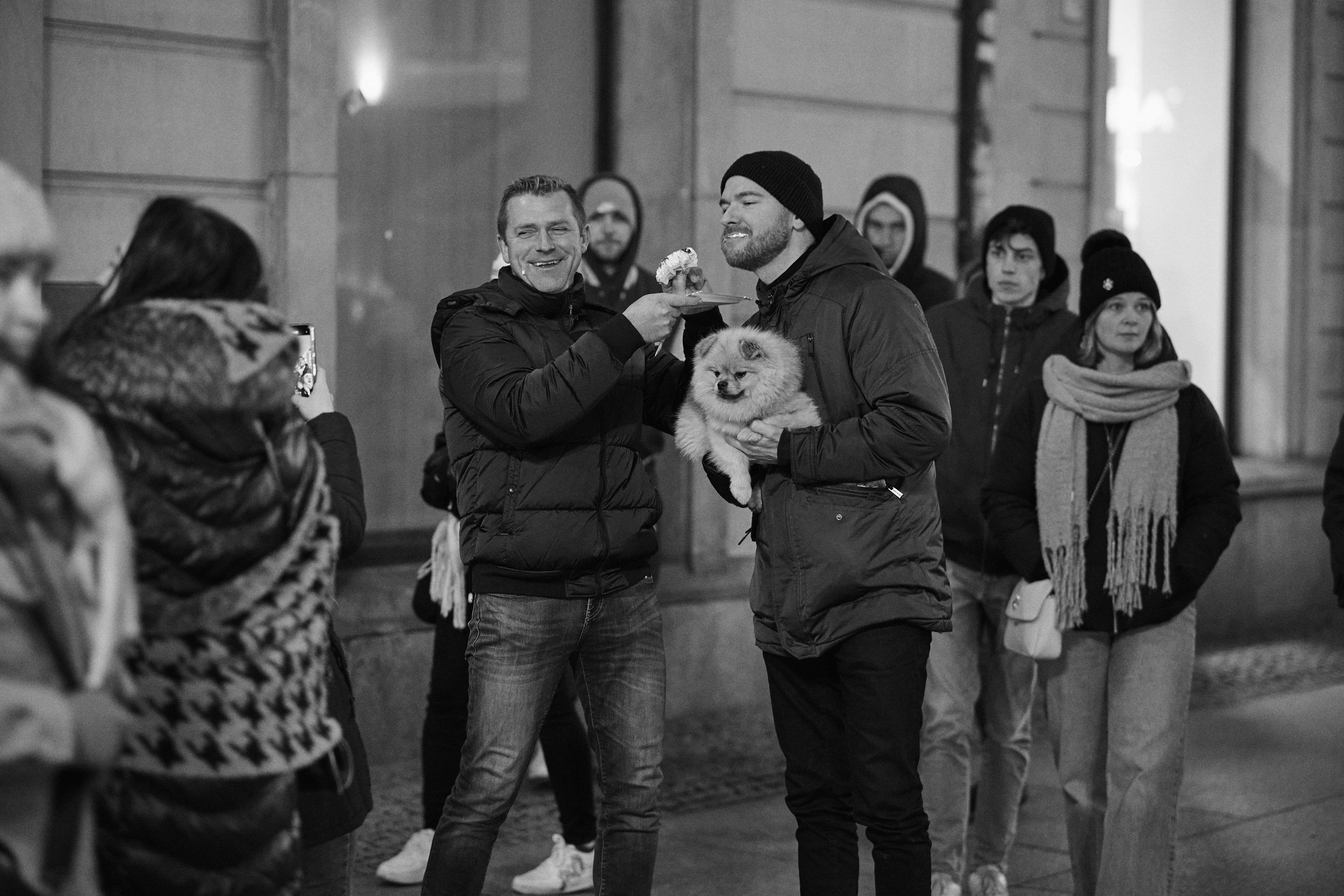 The image depicts a group of people standing casually on a city street. One person is holding a small fluffy dog. Another person is playfully holding something near the bearded man's face. The group is dressed warmly, suggesting a cold day or evening. The photo is in black and white.