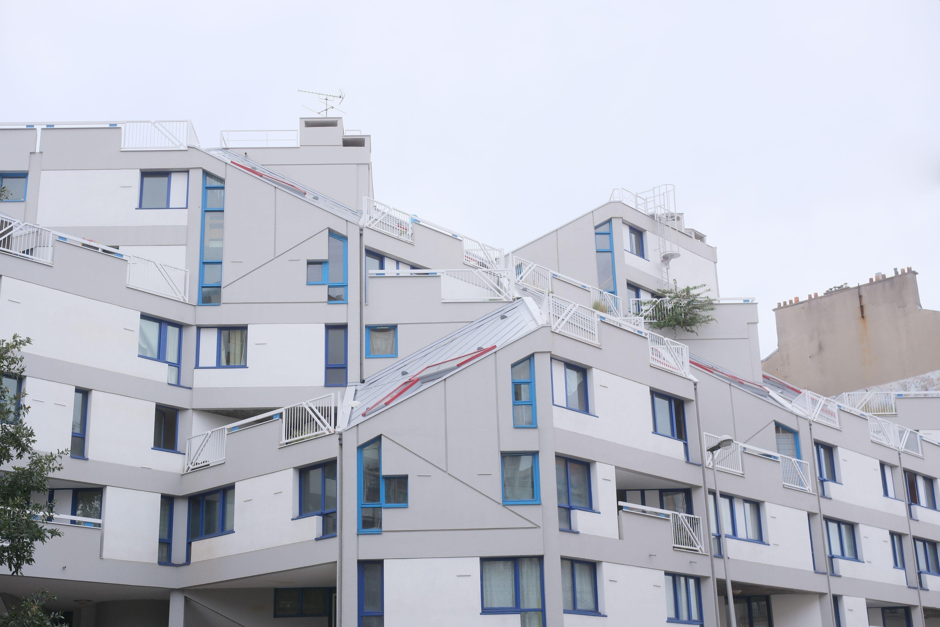 Photo horizontale. Un immeuble blanc dont l’architecture est un peu chaotique, tout en angles, des fenêtres de tailles et formes différentes et dont les bords sont bleu. Le tout sous un ciel gris