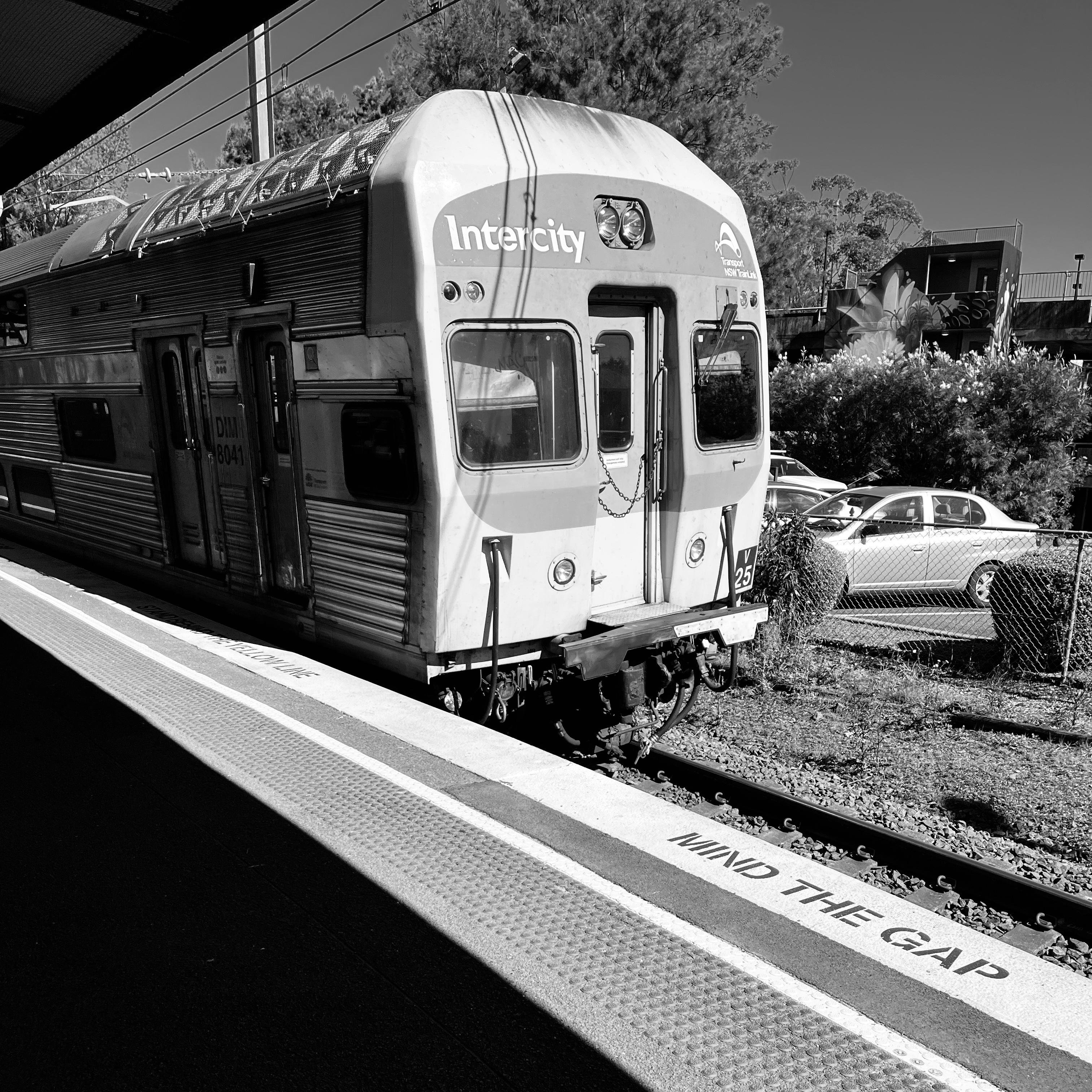 Black and white image of an intercity train stationary at a platform. The word "Intercity" is visible on the train's front. The platform edge has the warning "Mind the Gap" in bold letters. A few cars and trees are in the background, with shadows cast across the platform and tracks.