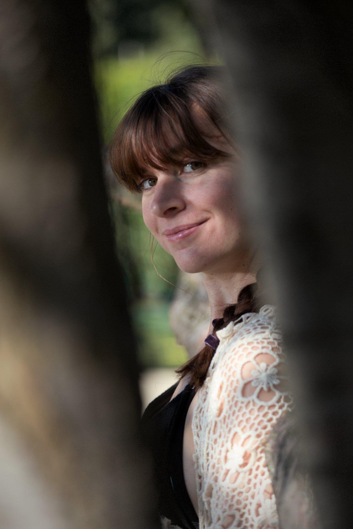 Photo portrait, montrant une jeune femme brune coiffée d’une natte pendant devant son épaule gauche, de profile, qui tourne la tête vers sa gauche pour regarder la caméra à travers 2 grosses branches d’arbres au premier plan, floues.