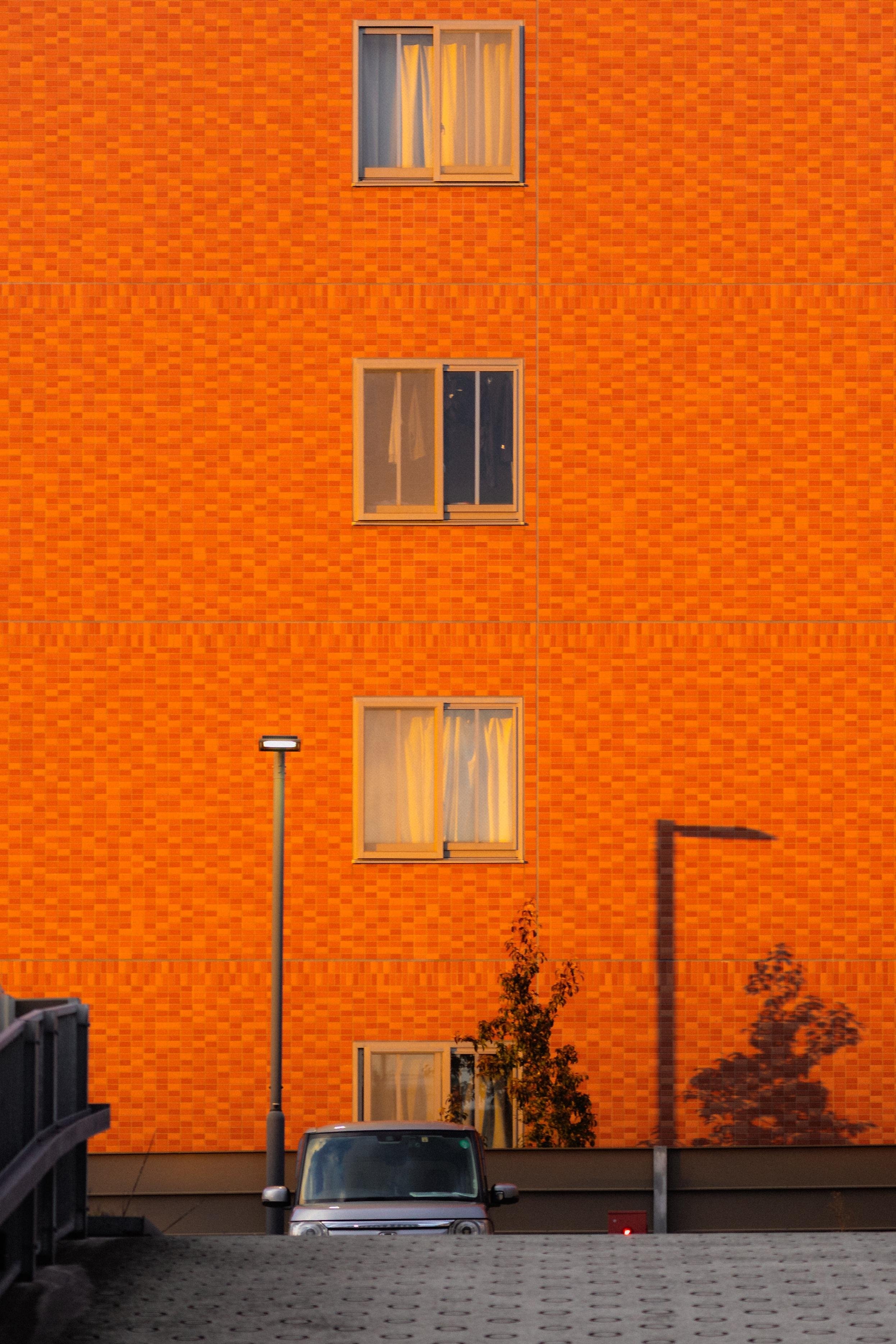 The image shows a vibrant orange building with a grid pattern. It has three windows, each with curtains. A car is parked below in front of the building, partially visible. There are shadows of a lamp post and a tree on the wall, indicating the direction of the light source. The setting appears to be a residential area, possibly late in the day with warm lighting.