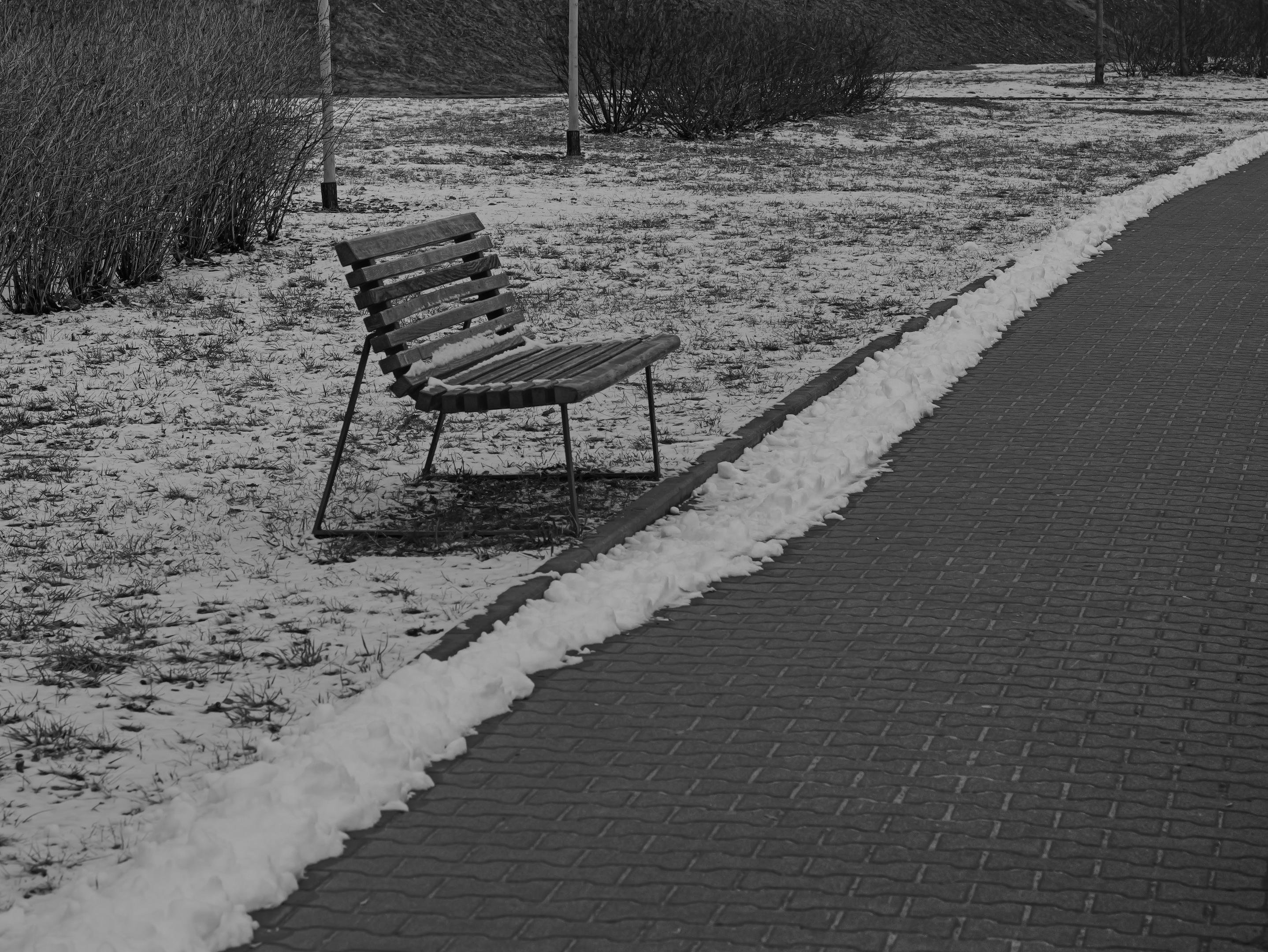 The image depicts an outdoor scene with a wooden bench on the left side. It is partially covered in snow, and the ground appears to be lightly dusted with snow as well. There is a paved path to the right, with snow outlining the edges, and some bushes in the background. The setting looks cold, suggesting a winter environment.
