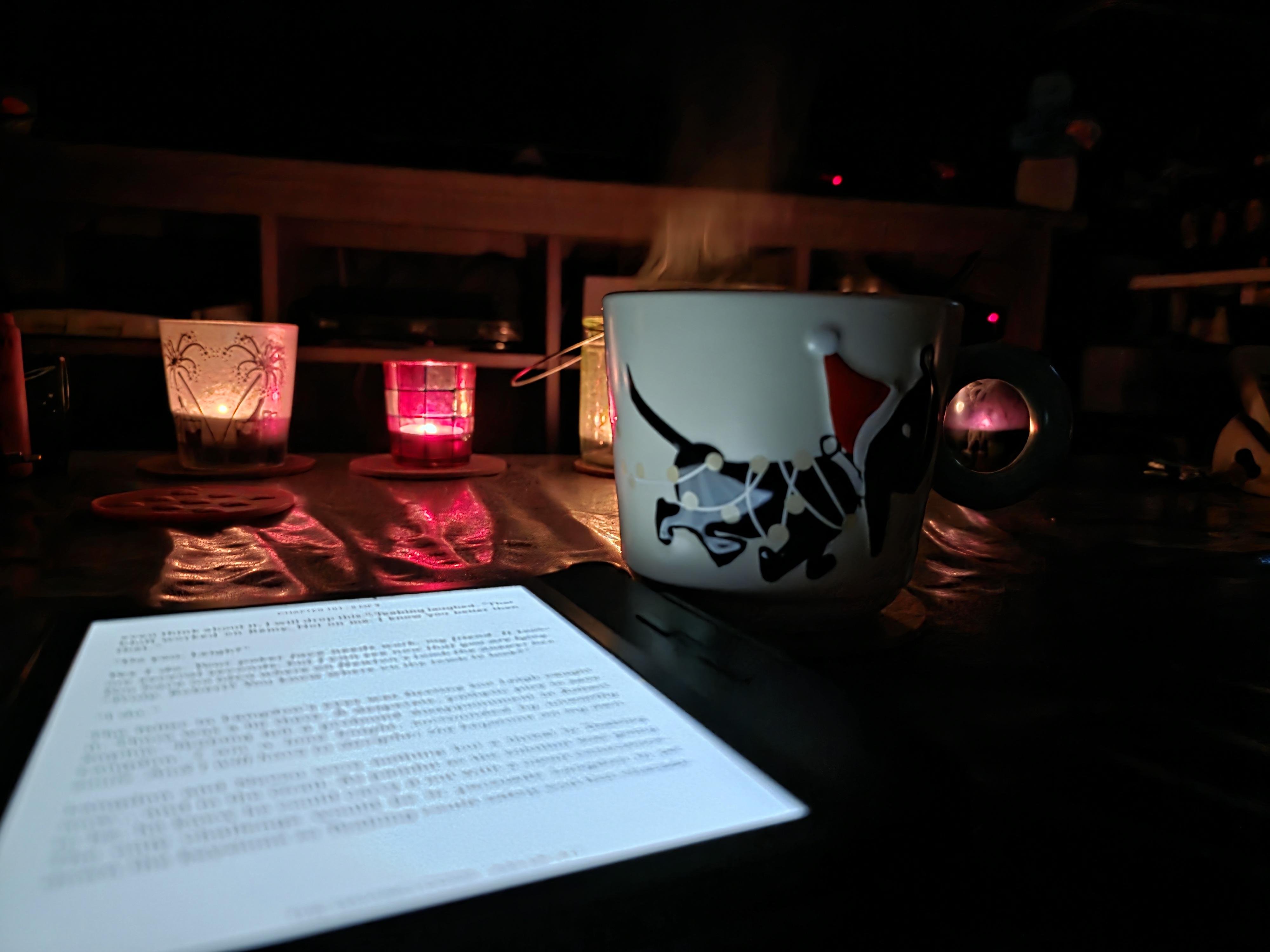 A darkened room, with several candles burning in colored glass holders. In the foreground to the left, lays a Kobo eReader, opened on a page of the DaVinci Code book, glowing bright in the darkness due to the back light settings. Next to the Kobo stands a big steaming mug. The mug has a black Dachshund on it, in Xmas style, based on drawings by Fiep Westendorp. 