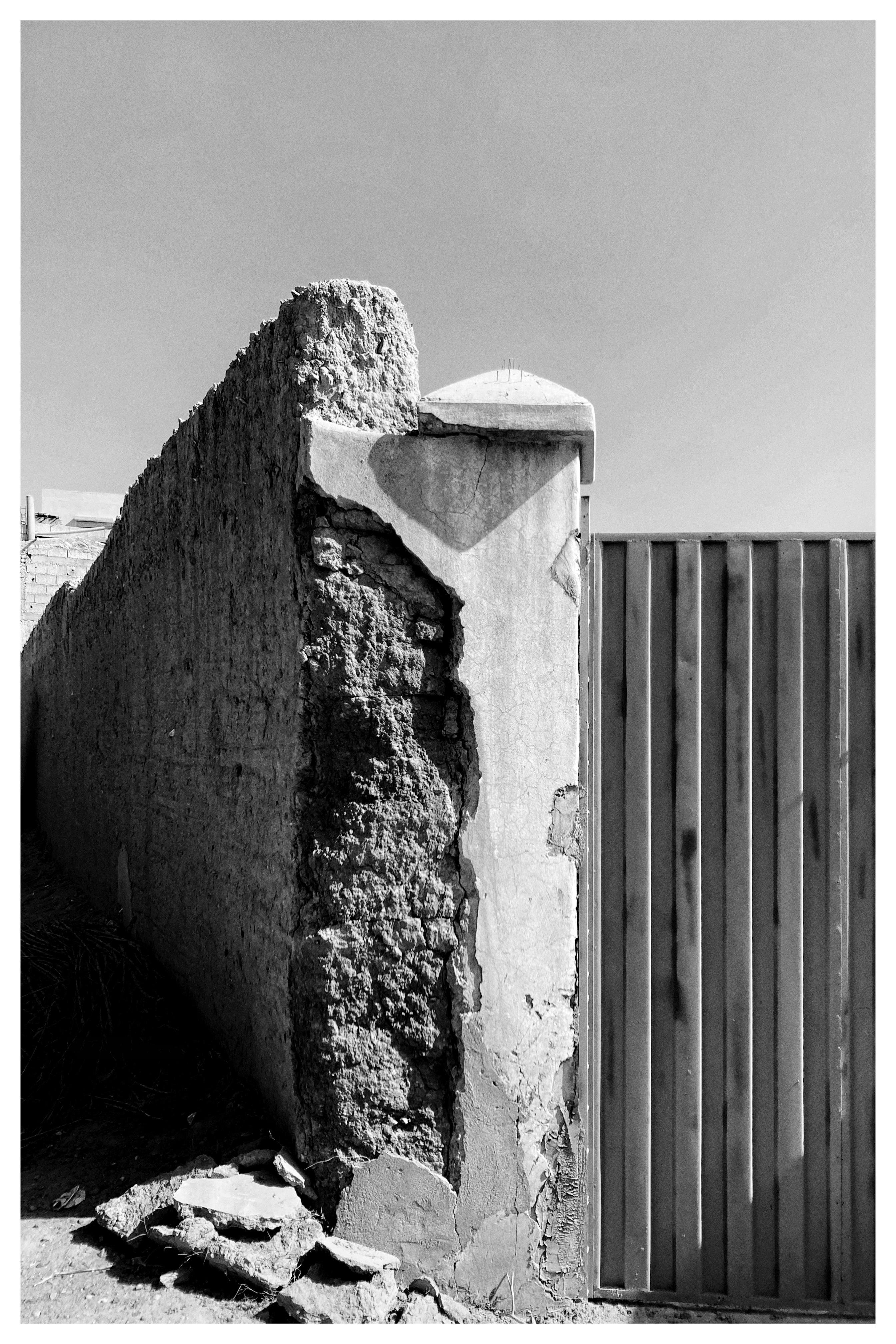 Black-and-white photo of a crumbling concrete wall and corner pillar with peeling plaster and rubble at the base, next to a tall corrugated metal gate under a clear sky.