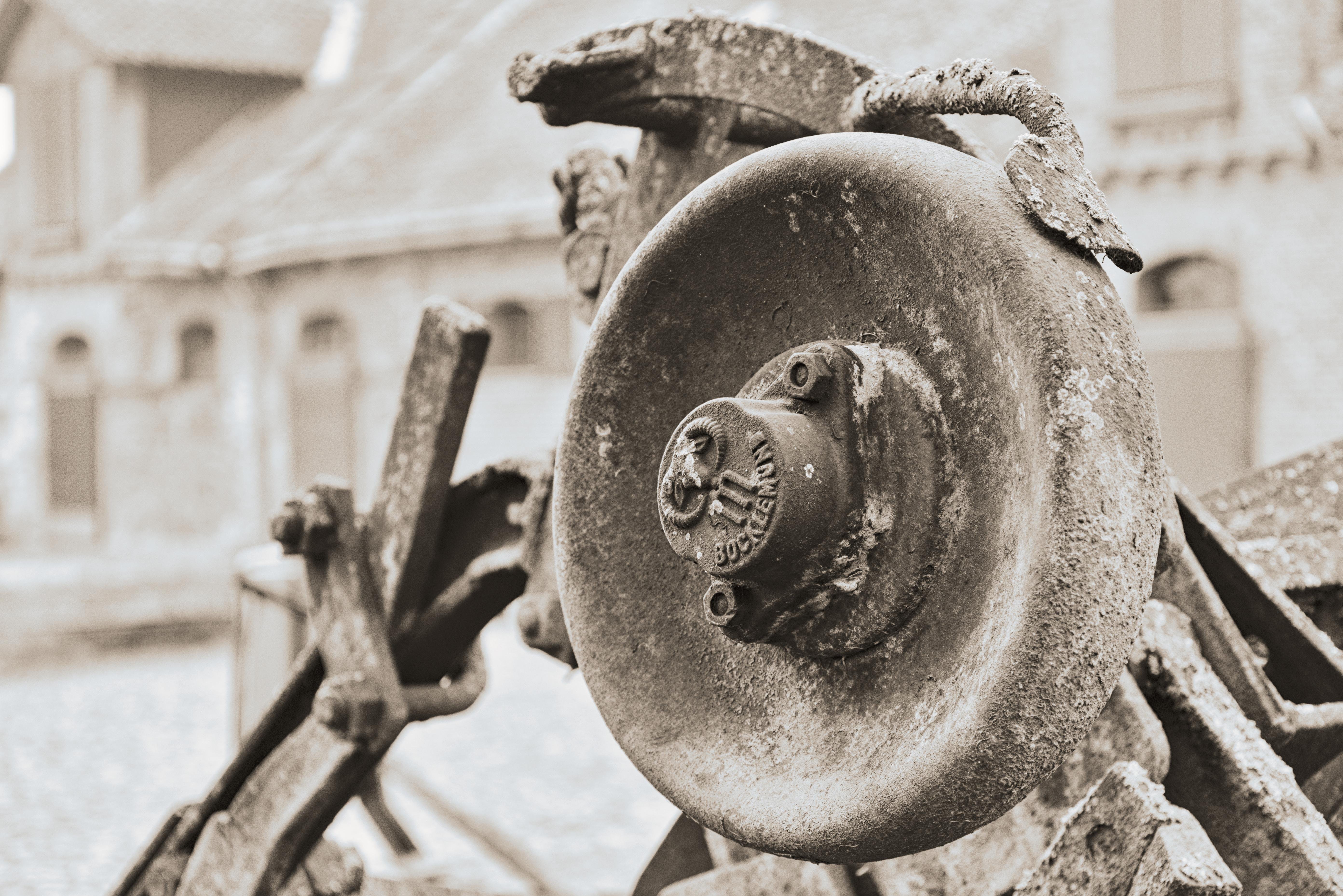 The image shows a close-up of an old, weathered gear or wheel mechanism, likely part of an industrial or agricultural machine. The metal appears corroded, with the surface showing signs of rust and wear. In the background, there is an out-of-focus building that looks like it might be part of an industrial site or an old structure. The photo is in black and white, giving it a vintage appearance.
