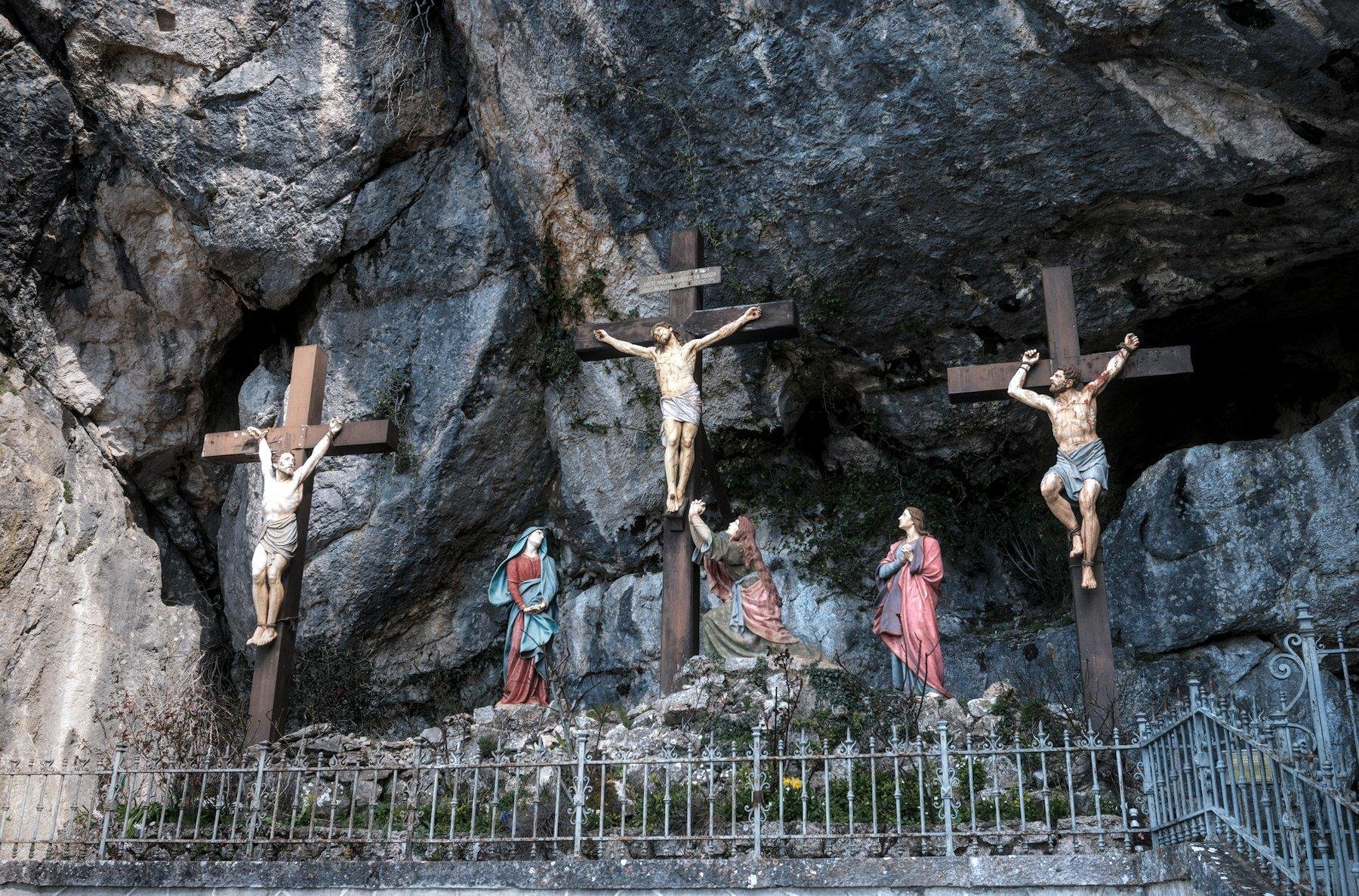 Photo horizontale. Des statues peintes représentant une scène biblique dans une alcove de la montagne. Au centre, Jésus crucifié, de chaque côté, deux hommes attachés à des croix qui regardent Jésus, 3 femmes au pieds de la croix de Jésus, priant. Une grille protège l’œuvre des touristes
