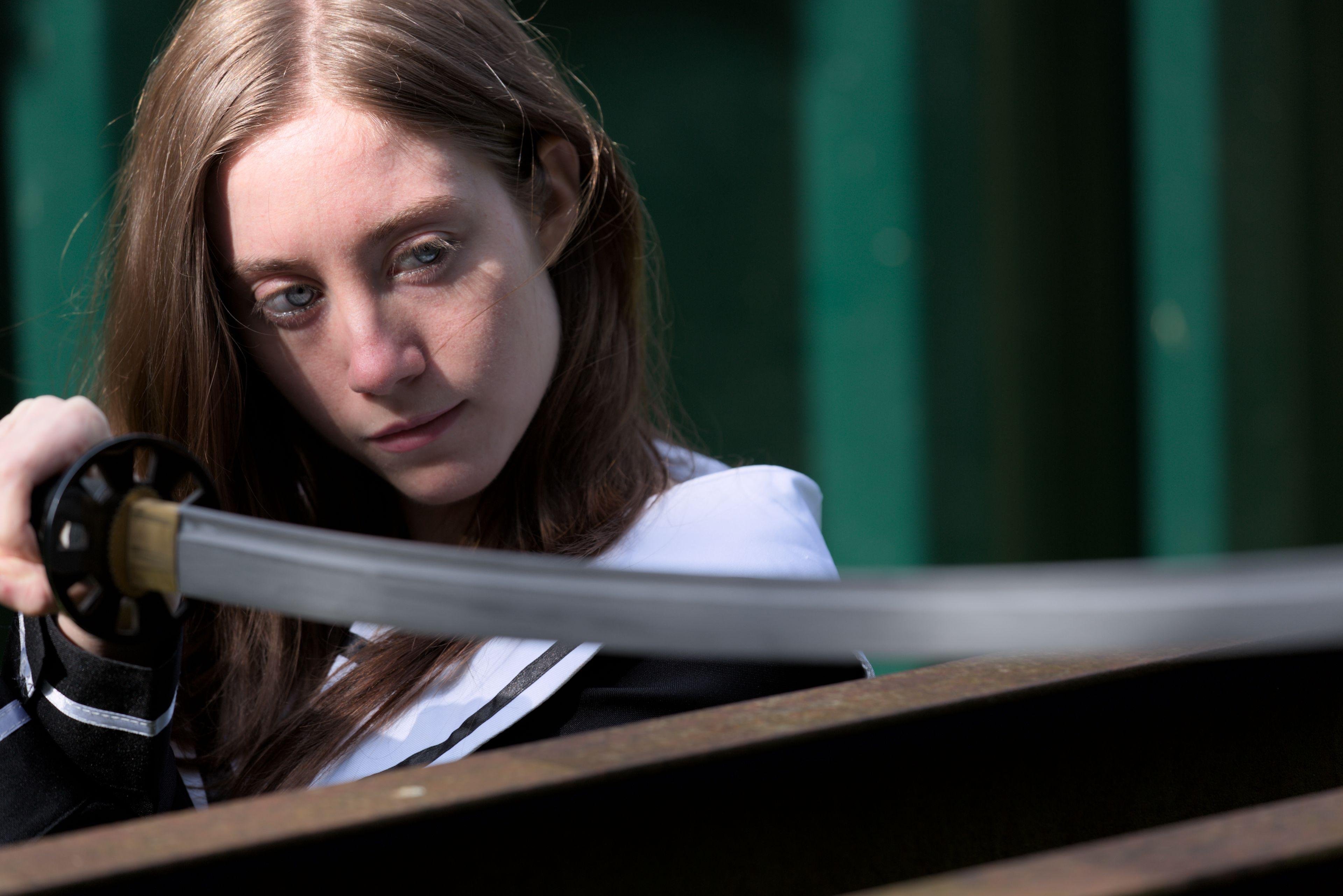 portrait of a woman in a japanese school uniform, holding a katana above a rusted steel beam, nearing her head to the blade to inspect it