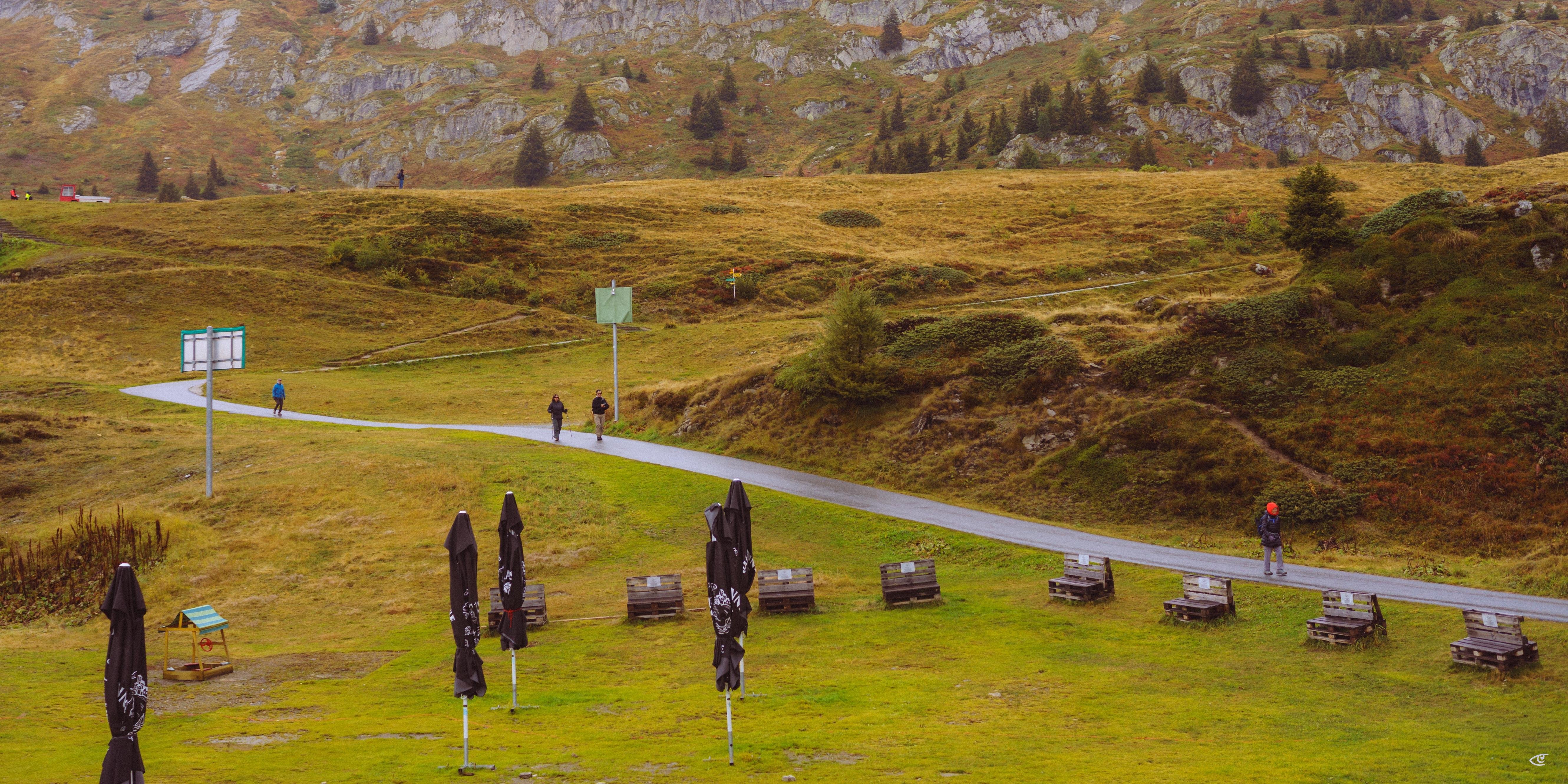 Mountain meadow with a paved path curving uphill, a few walkers on the path, and several closed black patio umbrellas with wooden picnic tables on the grass; rocky hillside with scattered trees in the background.