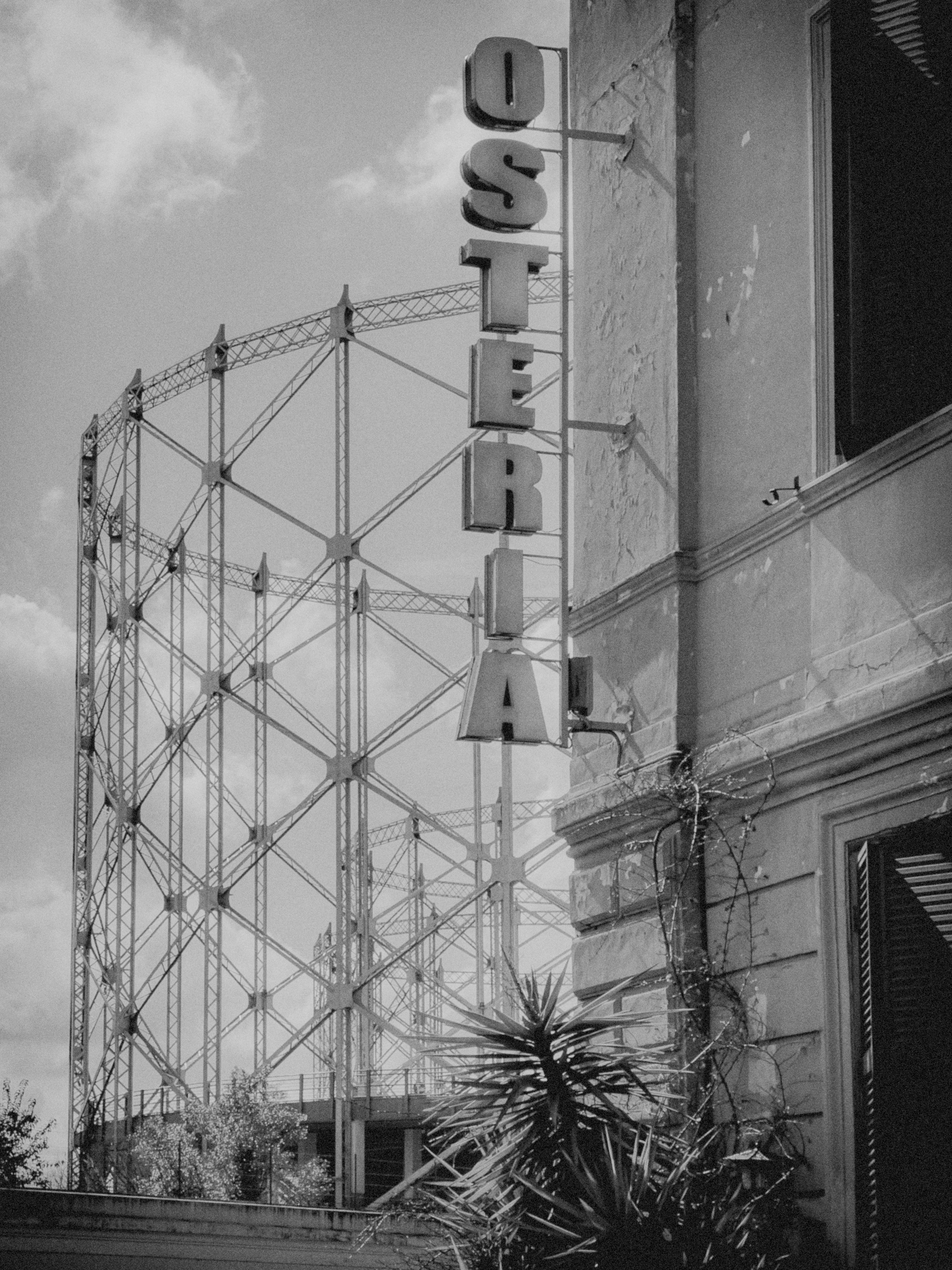 Struttura industriale, probabilmente un gasometro, sullo sfondo. In primo piano, c'è un edificio con un'insegna verticale che dice "OSTERIA." La scena è in bianco e nero, con della vegetazione visibile vicino all'edificio.