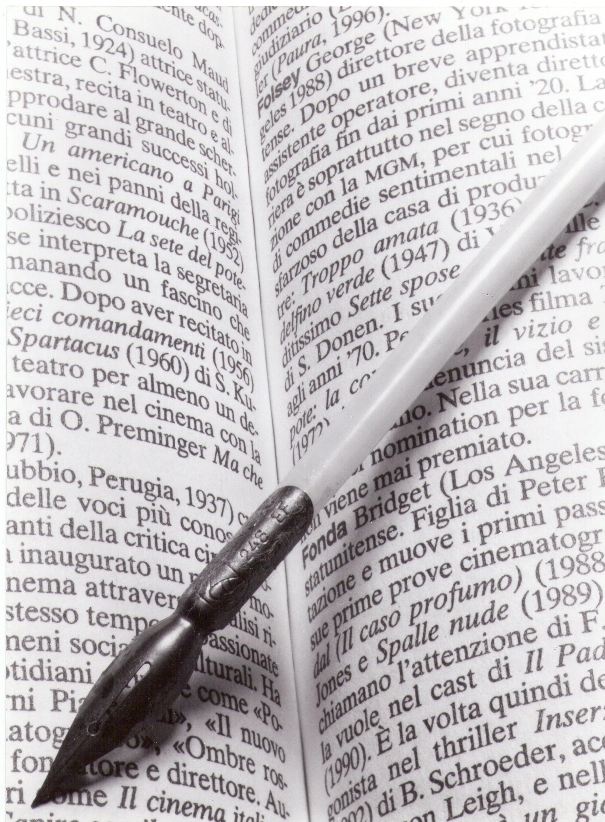The image shows an open book with text in Italian. There is a calligraphy pen resting on the book's pages. The pen has a metal nib and a white handle.