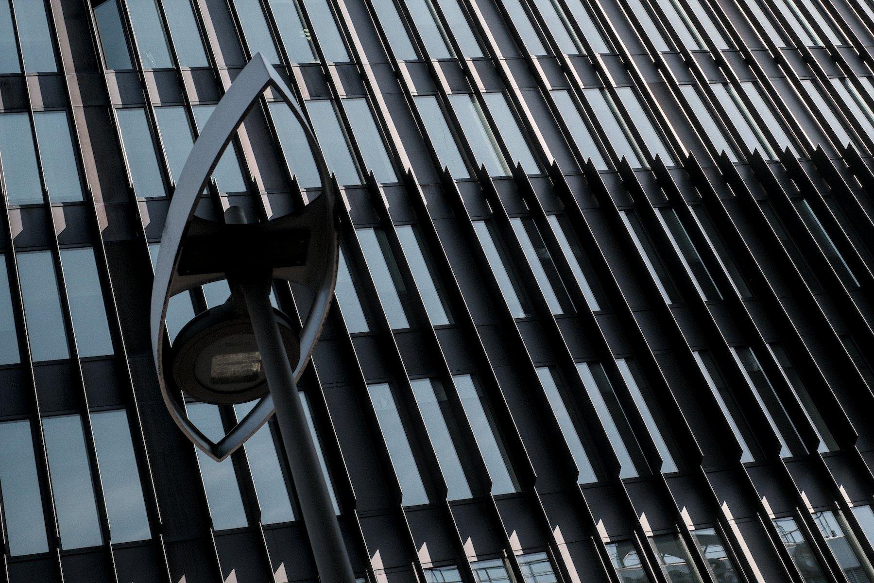Photo horizontale. Au premier plan à gauche, le haut d’un lampadaire au design moderne en forme de navette faisant penser aux calissons d’Aix. En arrière plan, la façade d’un bâtiment moderne en verre et acier, strié de barres métalliques noires verticales.