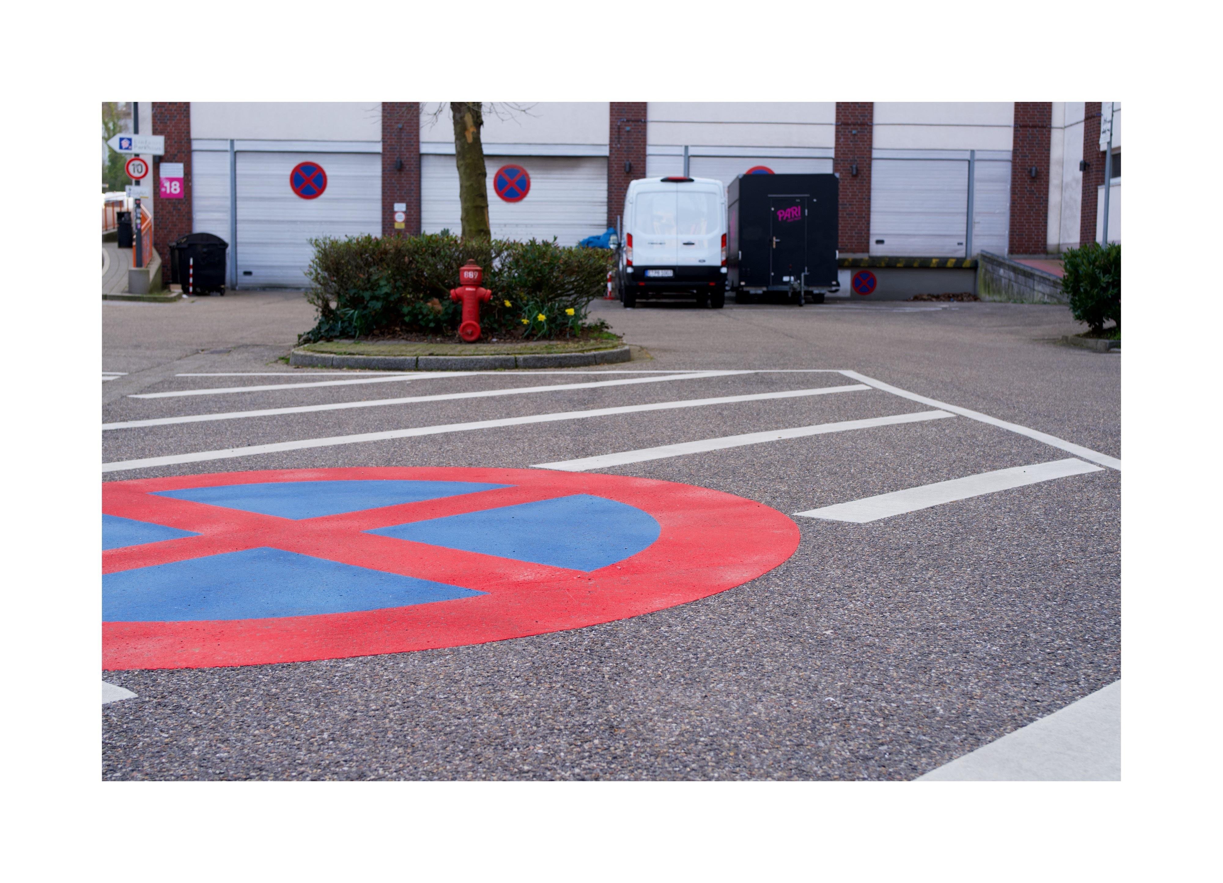 Ein Parkplatz. Auf dem Boden ist ein großes Parkverbotssymbol als blaues Schild mit rotem Kreuz und rotem Rand aufgemalt, darum herum ist in weißen Streifen eine Sperrfläche markiert. Im Zentrum des Bildes steht ein roter Hydrant in einem kleinem, grünen Beet. Im Hintergrund sind Rolltore zu sehen, auf denen sich das Parkverbotssymbol mehrere Male wiederholt.