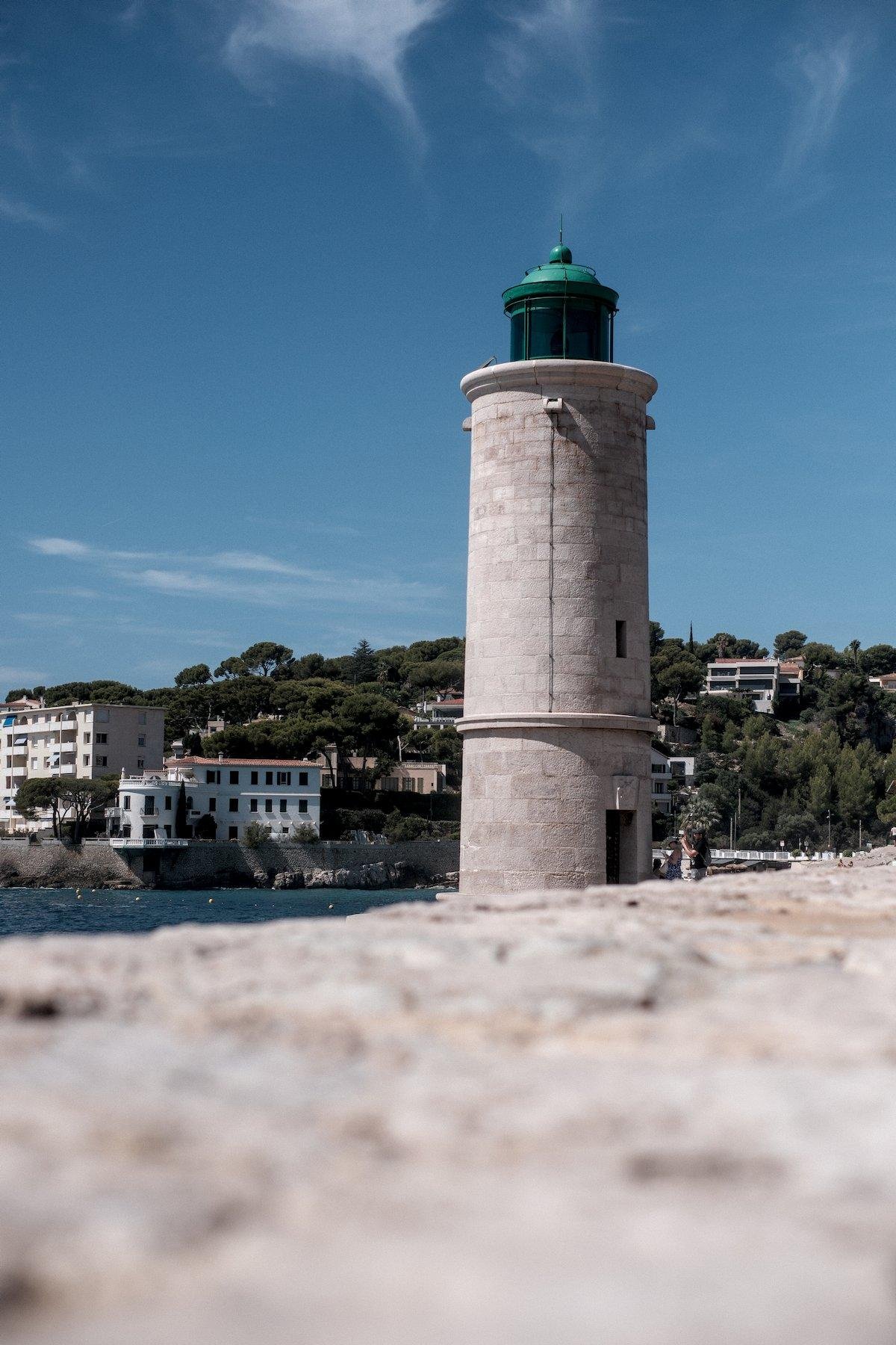 Vertical photo. The foreground is a low level view of a stone wall, blurred by the depth of field. The wall leads the eye to a lighthouse, half lighted by the sun from the left. It is made of the same clear coloured stone as the wall and the top is painted in green. At the base, on the left, a bit of water from the sea, and behind, some habitation building with trees among them, below a blue sky with a dash of light clouds