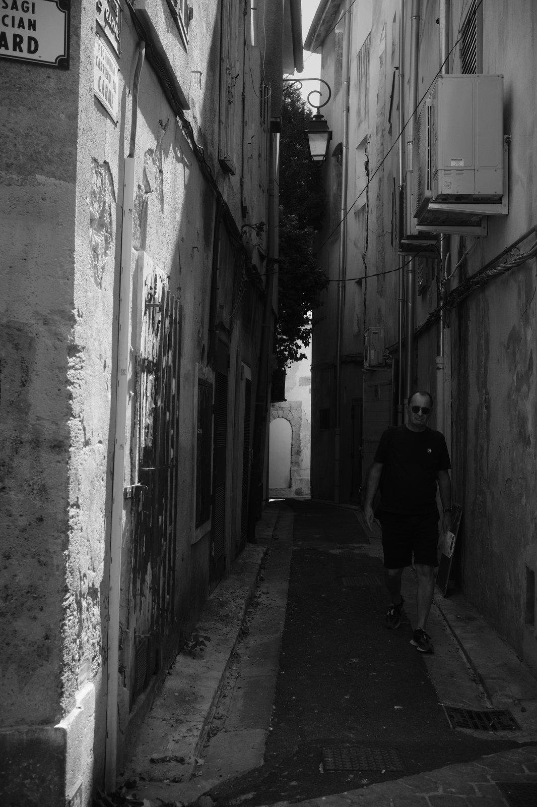 Black and white photo of a small dark alley from which a man with shades is walking out. Some sun lights the beginning of the alley and we can see some light at its end too. An air con exterior unit is above the man.