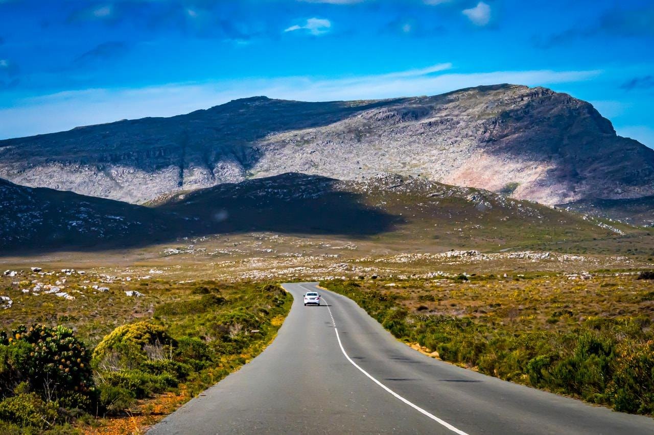 The image depicts a scenic landscape with a straight road leading toward a large, rocky mountain. There are a few cars traveling on the road, surrounded by green, grassy terrain and under a bright blue sky.