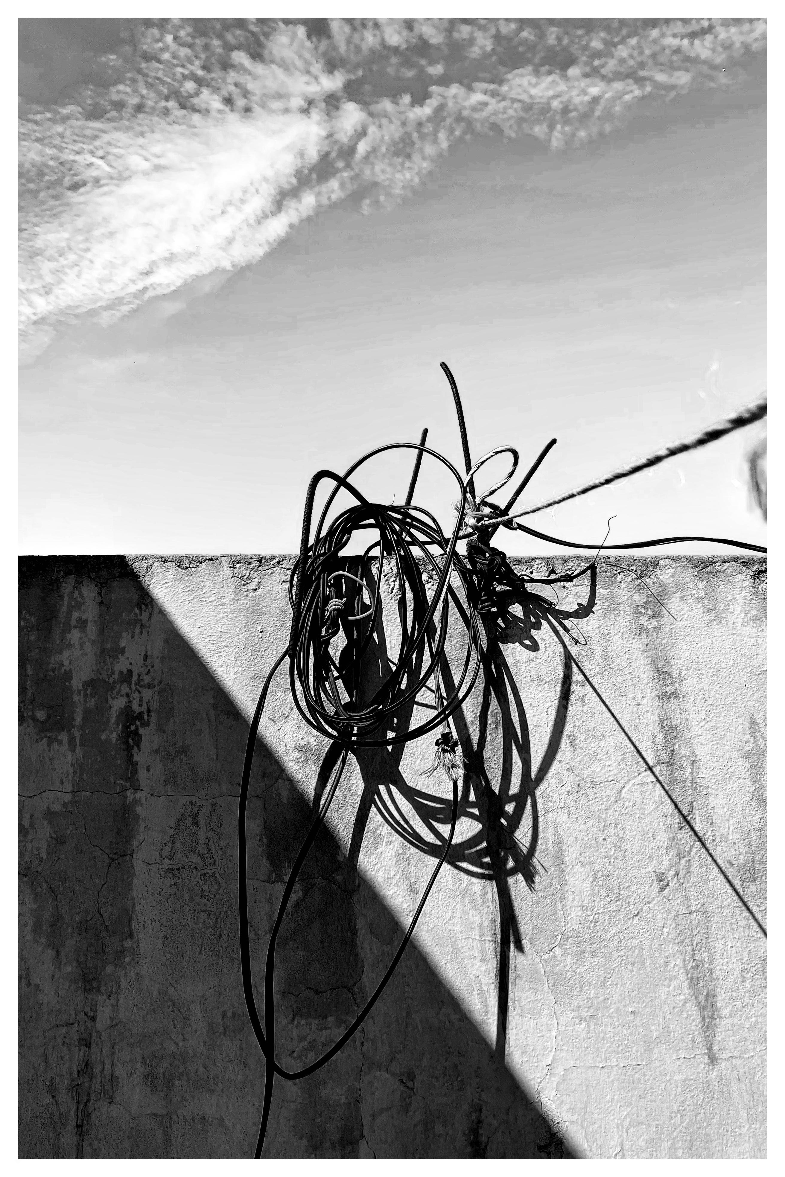 Black-and-white photo of a weathered concrete wall under a cloudy sky, with a tangled coil of cables draped over the top edge and their shadows cast diagonally across the wall.