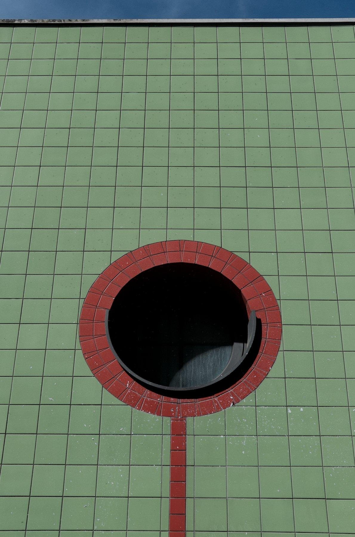 Photo verticale. Un mur couvert de carrelage vert, avec une fenêtre ronde, cerclée de carrelage rouge et une ligne de carrelage rouge qui descend. Un bout de ciel bleu tout en haut de la photo