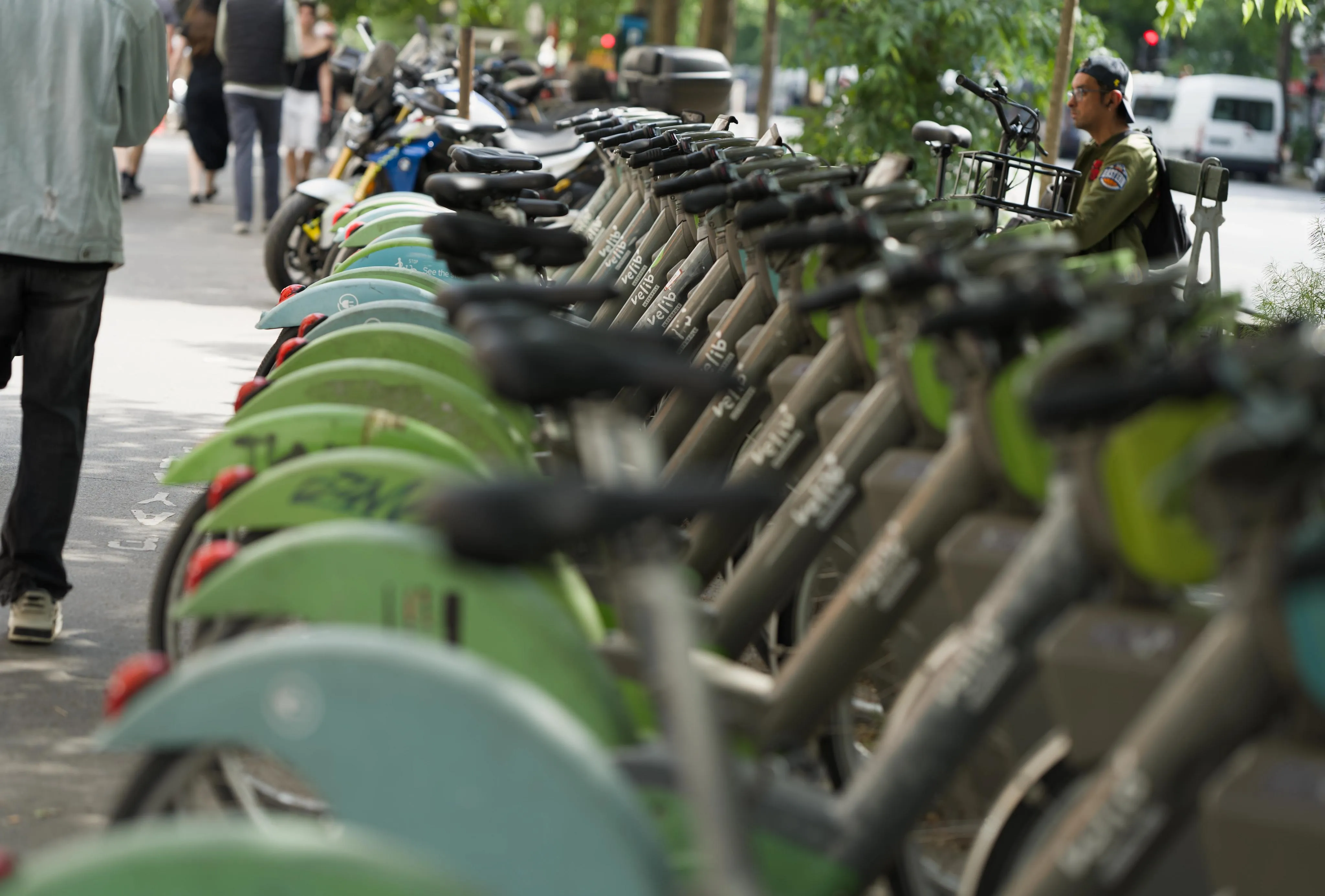 Daytime photograph of multiple public use bicycles stationed on a street sidewalk. The stationed bicycles are forming a row, taken from the side at the end of the row. Most bicycles have green casing under their handle and over the rear wheel. A few of the bicycles have a turquoise casing in the same places. In the background behind the bicycles, a man wearing green clothing is sitting on a bench with a different model of bicycle stationed leaning against him, outside of the row of public use bikes.
