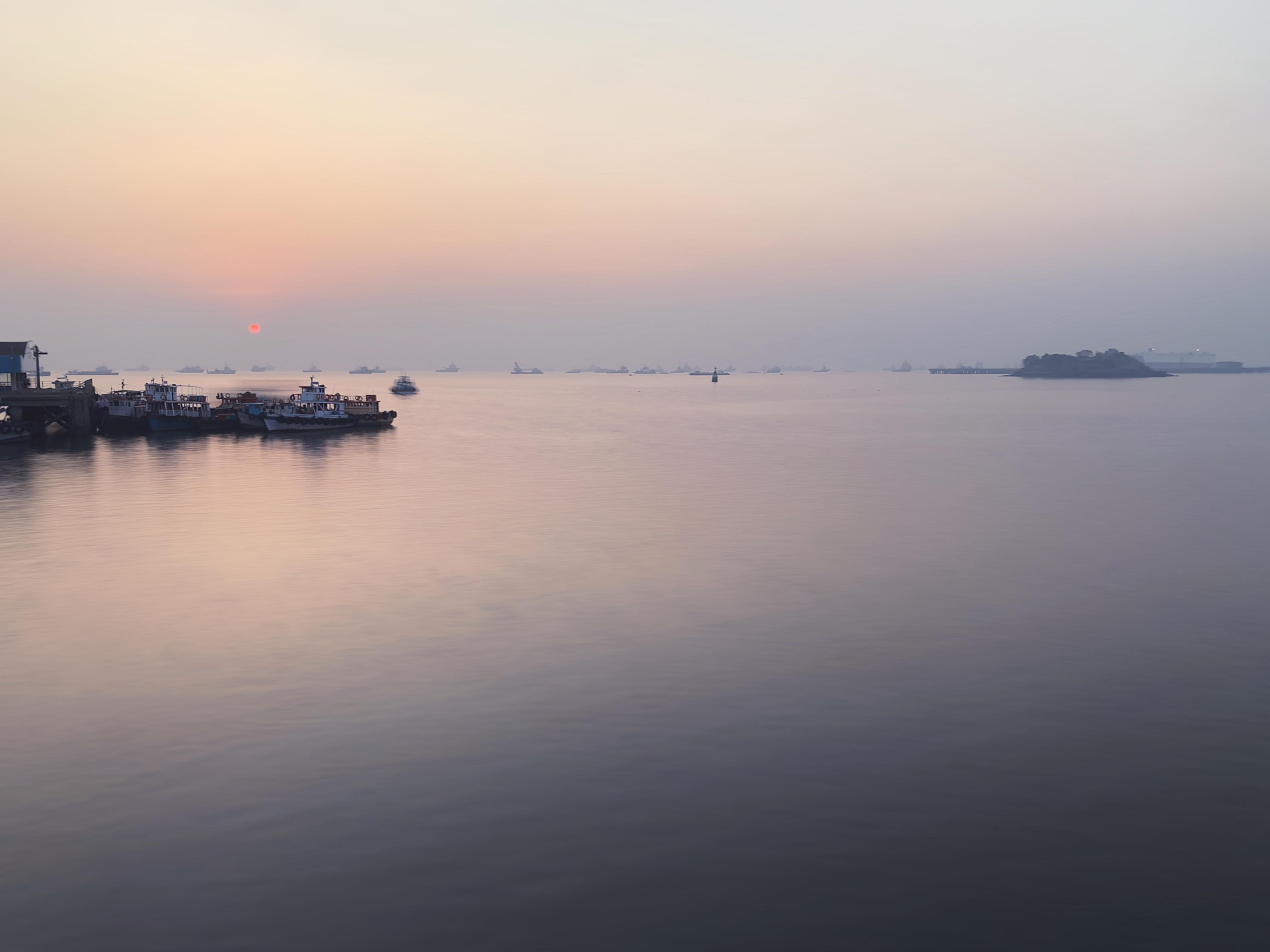 The image shows a tranquil seascape during sunset, with a calm water surface reflecting the soft colors of the sky. There are some boats docked on the left side, and a small island fort is visible in the distance to the right. The overall atmosphere is peaceful and serene.