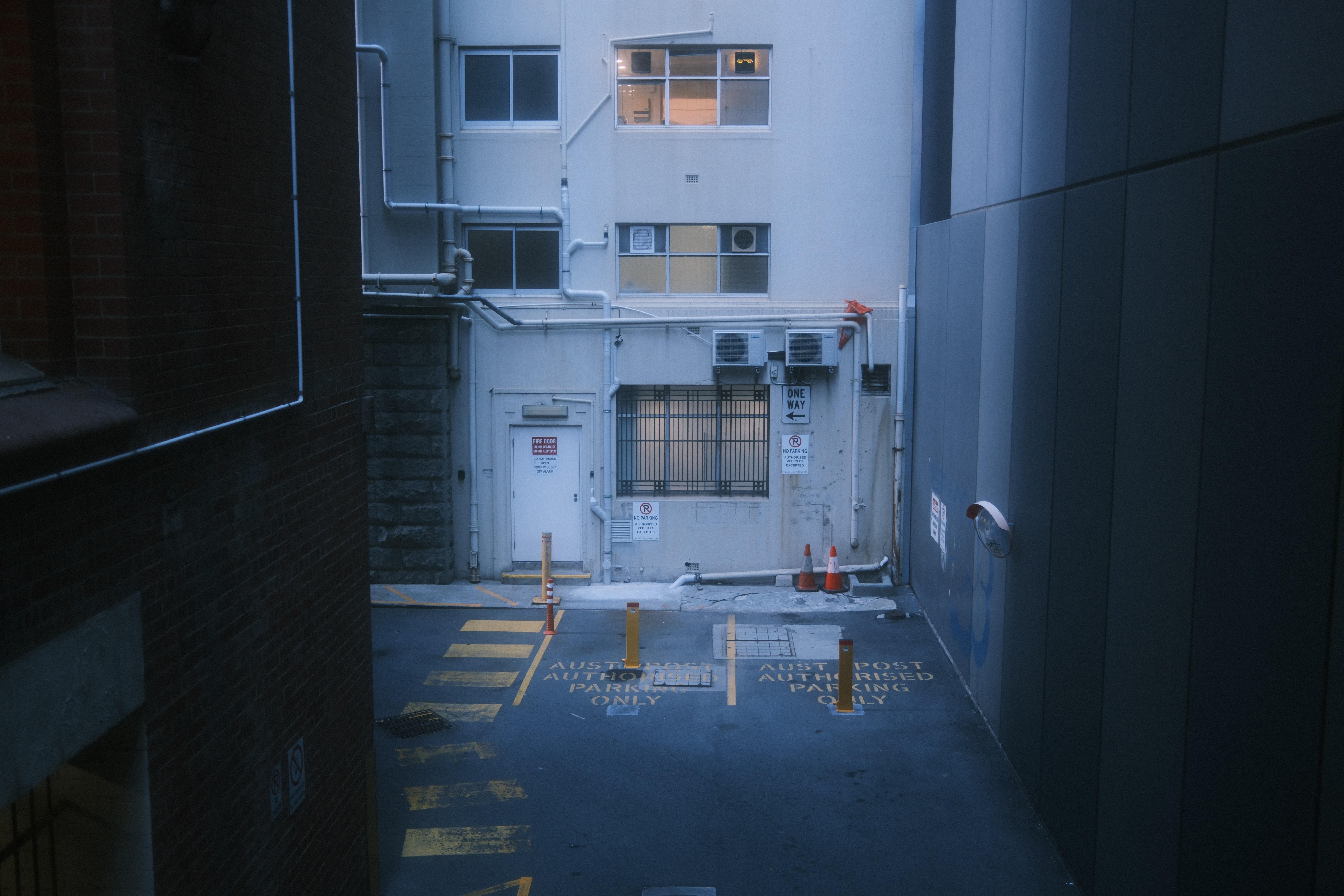 Narrow urban alley between tall buildings leading to a loading area, with painted lane markings, several yellow bollards, a barred window, wall-mounted air-conditioning units, a one-way sign, and two orange traffic cones near a closed door.