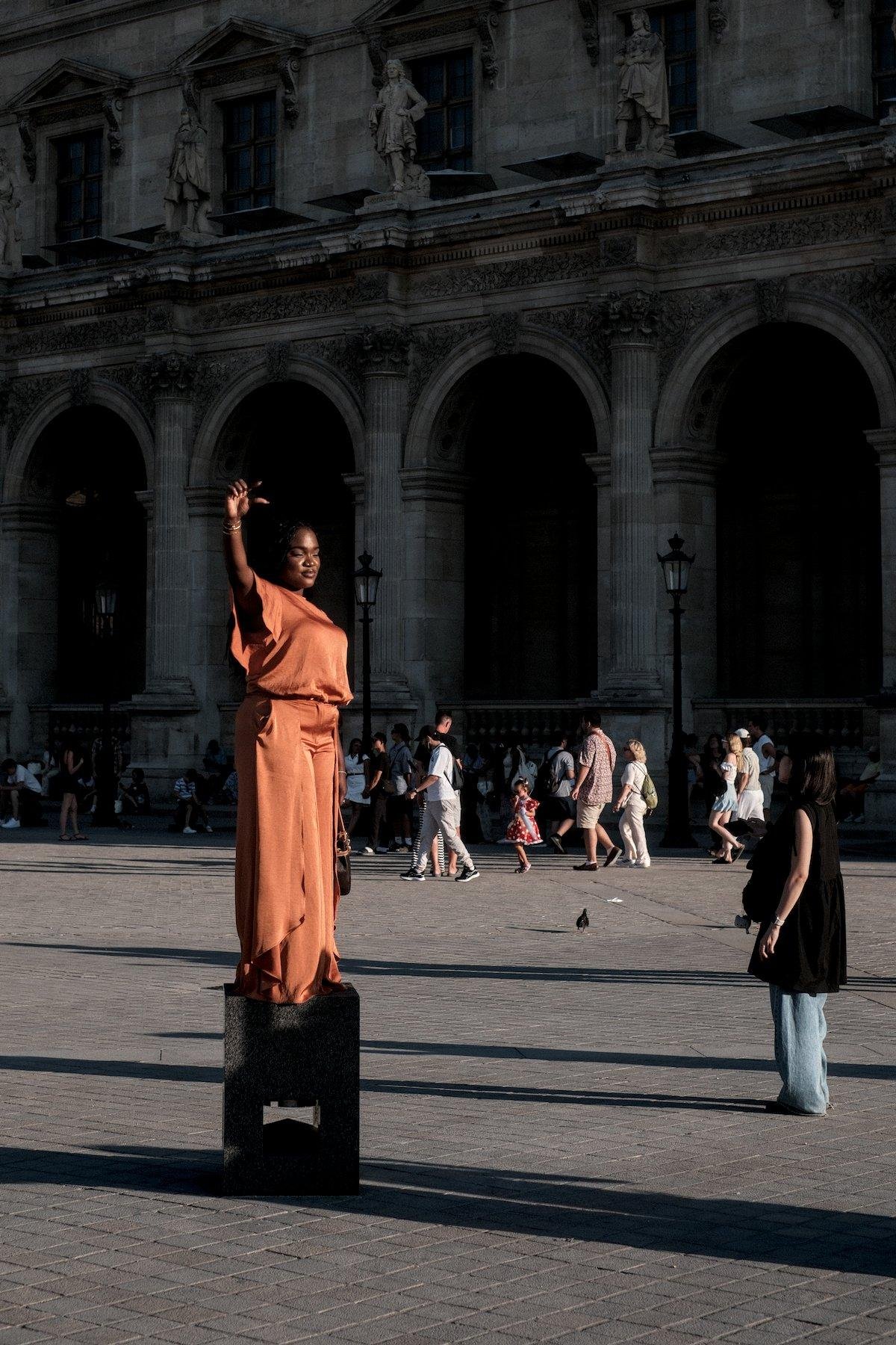 Photo verticale. Sur le parvis du Louvre, une femme en robe orange est perchée sur un petit piédestal et prends la pose faisant mine de pincer la pyramide du Louvre (hors champs à gauche). Le soleil couchant l’éclaire généreusement