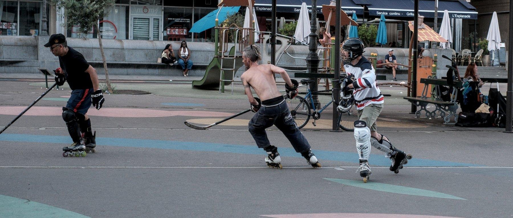 Photo horizontale format cinémascope. 3 hommes sur patins entrain de jouer à du hockey sur bitume. L’un d’eux est avec des pièces d’armures de hockey aux tibia et un casque