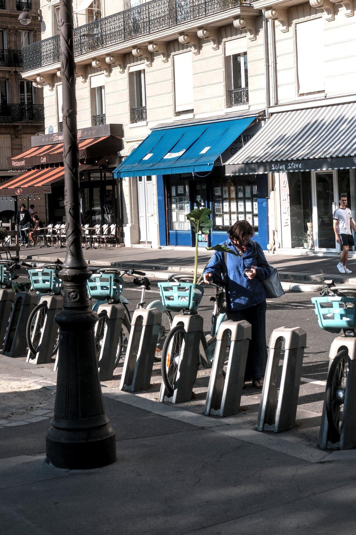 Photo verticale. Une jeune femme brune cheveux courts, blouson bleu, aux côté d’un vélib dans lequel une plante verte est posée. Elle est assez haute et dépasse la femme qui est en train de consulter son téléphone.