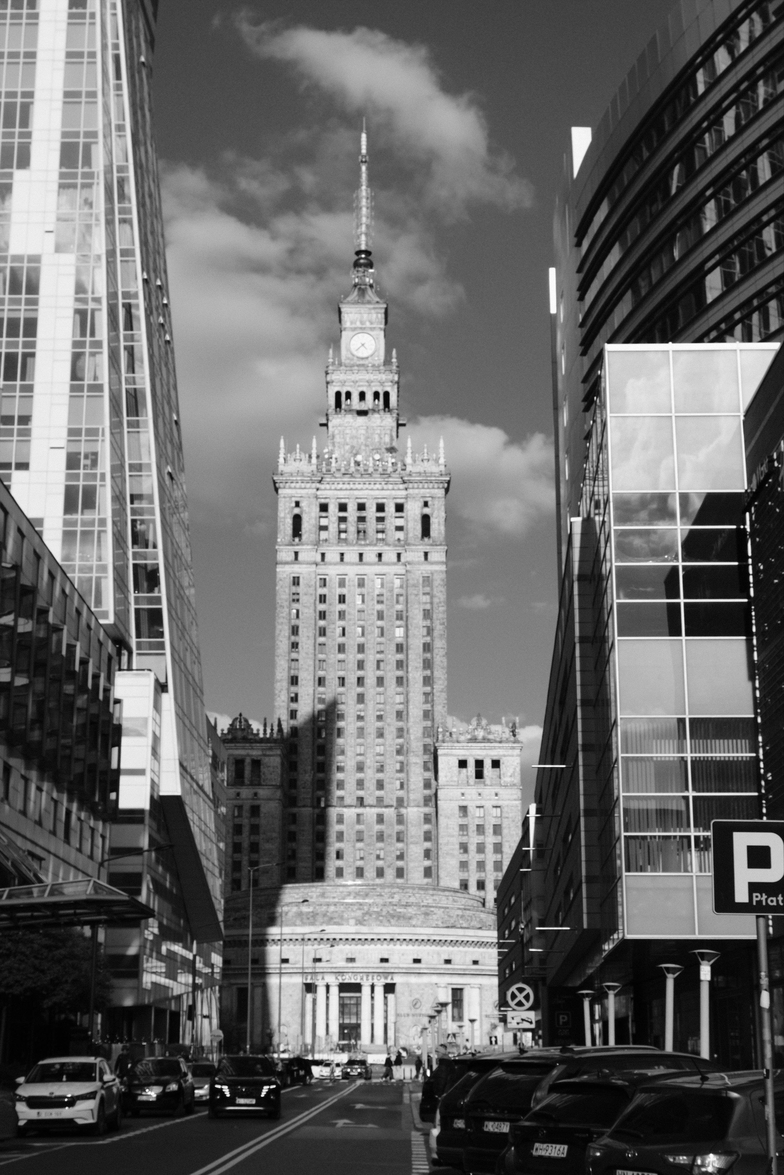 View along a city street onto the Palace of Culture and Science in Warsaw, a monumental building in the style of Socialist Classicism. On both sides modern office buildings and parked cars contrast the classicist architecture of the tower.