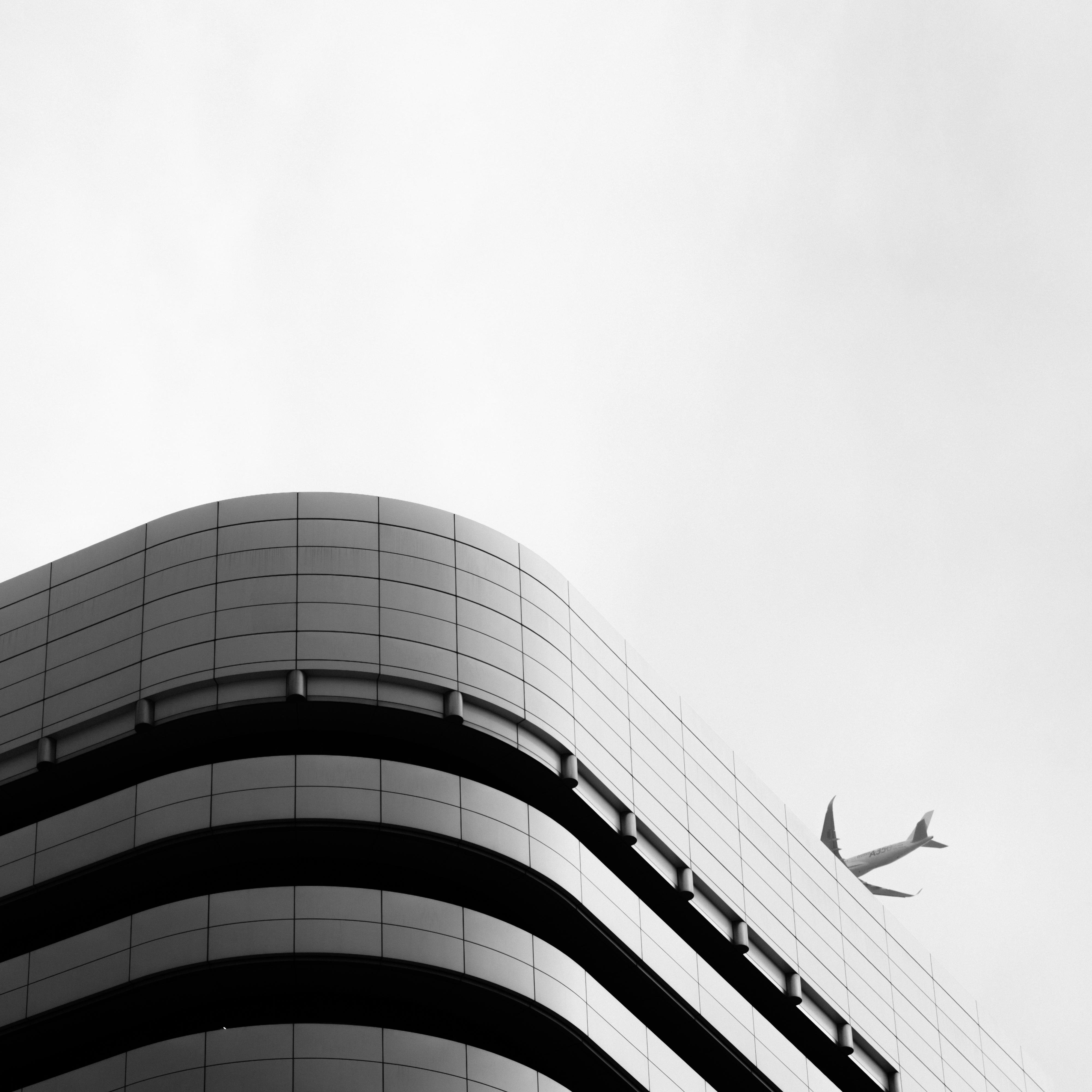 Curved corner of a modern building with a grid-like facade in the foreground, set against a cloudy sky. An airplane is flying in the distance above the building. The image is in black and white.