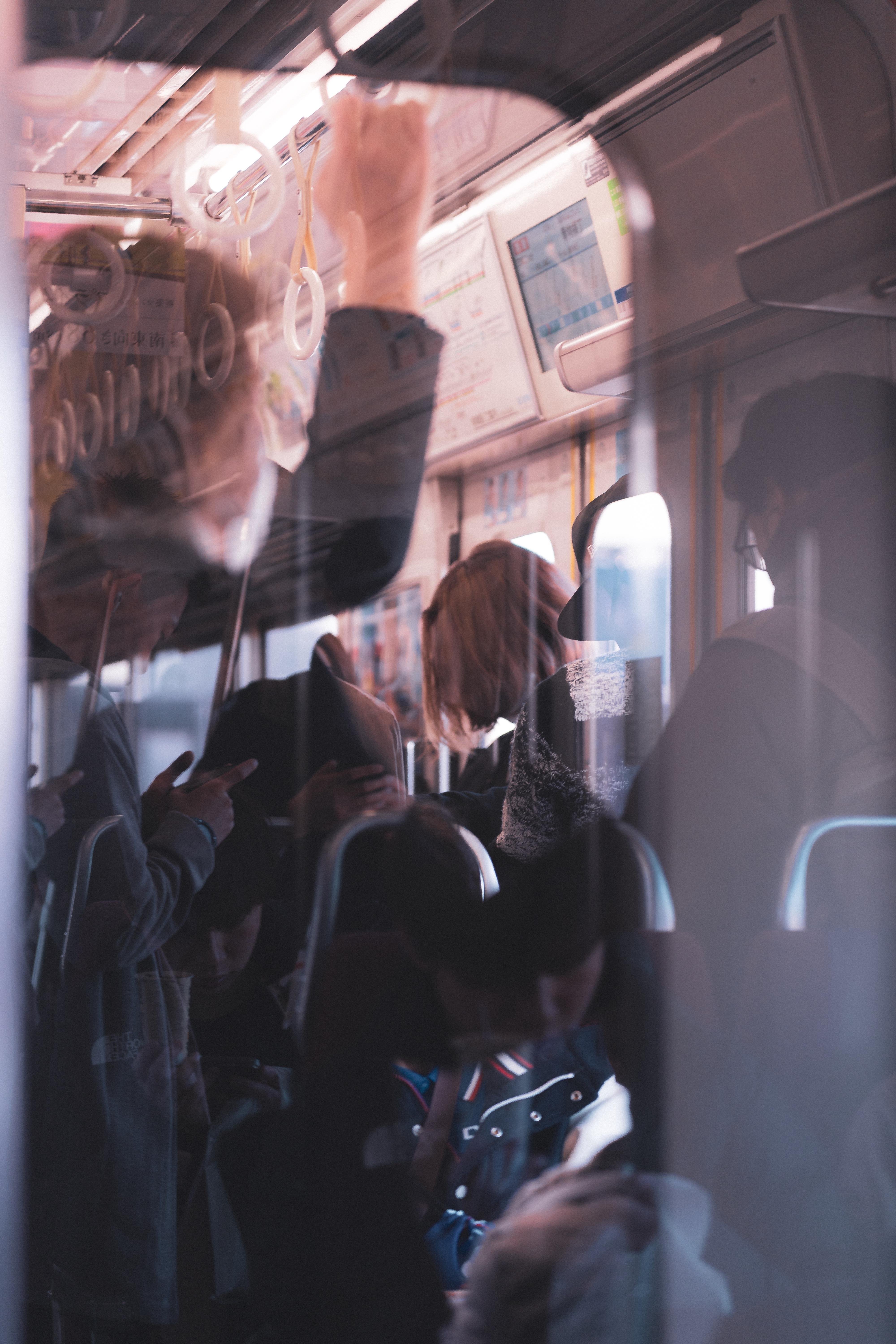 The image depicts a crowded setting inside a train or subway. People are standing and holding onto hand straps, with some looking at their phones. The image reflects a busy commute, emphasizing the close proximity of the passengers. The scene is dimly lit, adding a slightly reflective quality, possibly due to a window or glass partition.