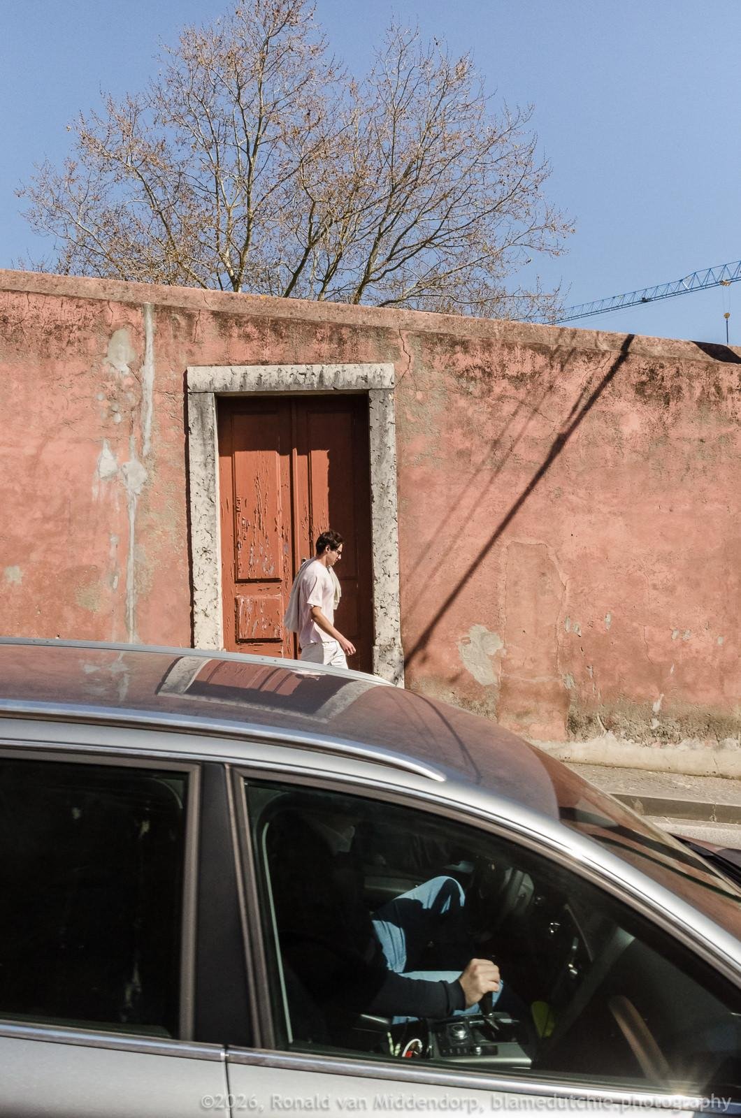 Man walking past a weathered pink stucco wall with a faded brown double door, while a dark car passes in the foreground; leafless tree branches and a clear blue sky above.
